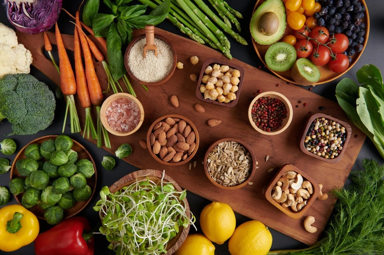 Various fresh vegetables, fruits, nuts, seeds, and spices arranged on a wooden board and surrounding bowls. Includes carrots, cauliflower, broccoli, Brussels sprouts, bell peppers, tomatoes, avocados, kiwi, grapes, lemon, and leafy greens, along with bowls of spices and nuts.