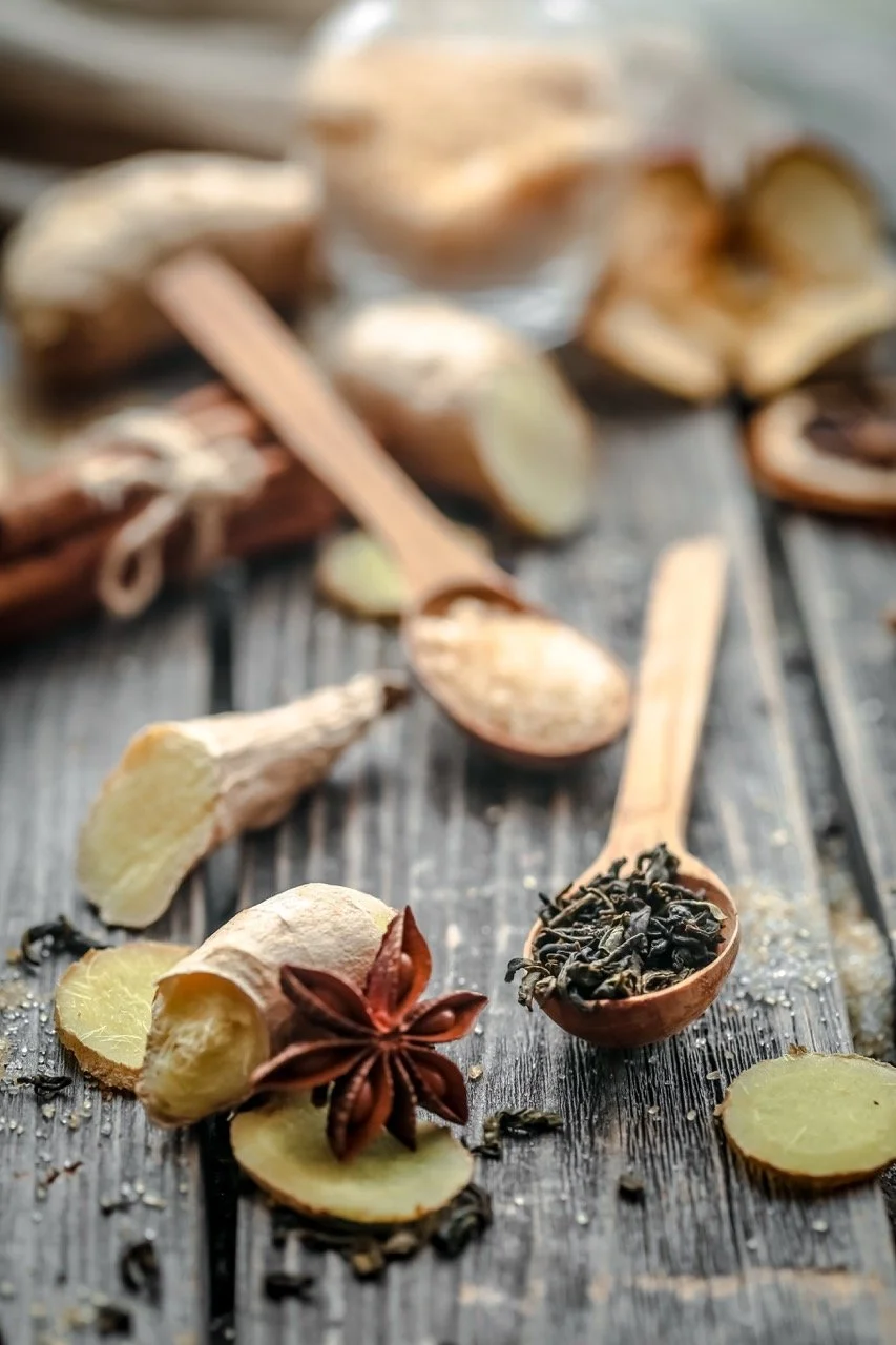 Several slices of ginger, a star anise, and loose black tea leaves on a wooden surface with a wooden spoon containing tea leaves in the background.