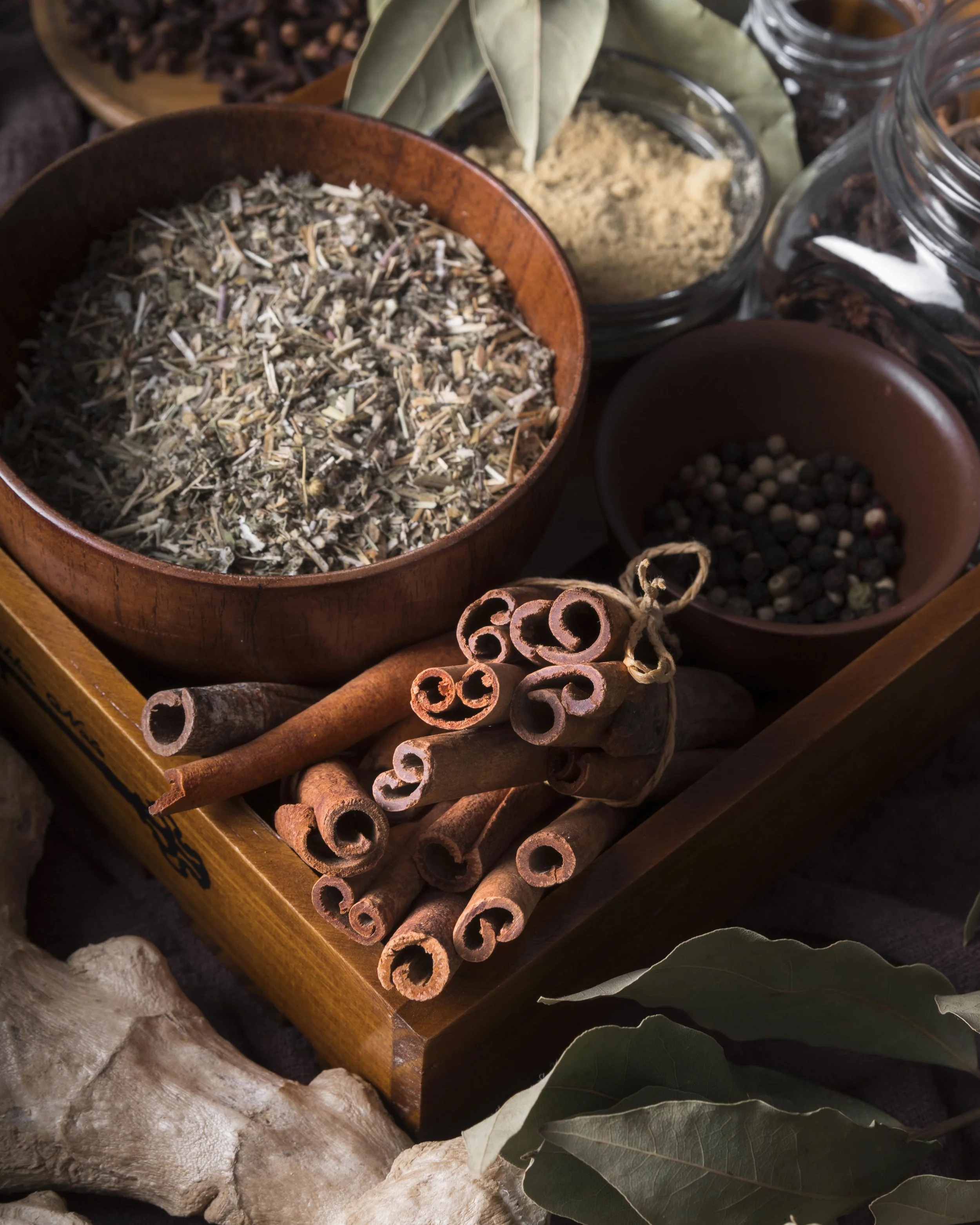 A collection of dried herbs and spices, including cinnamon sticks, bay leaves, and various ground spices in glass containers, arranged on a wooden tray with rocks and large leaves surrounding them.