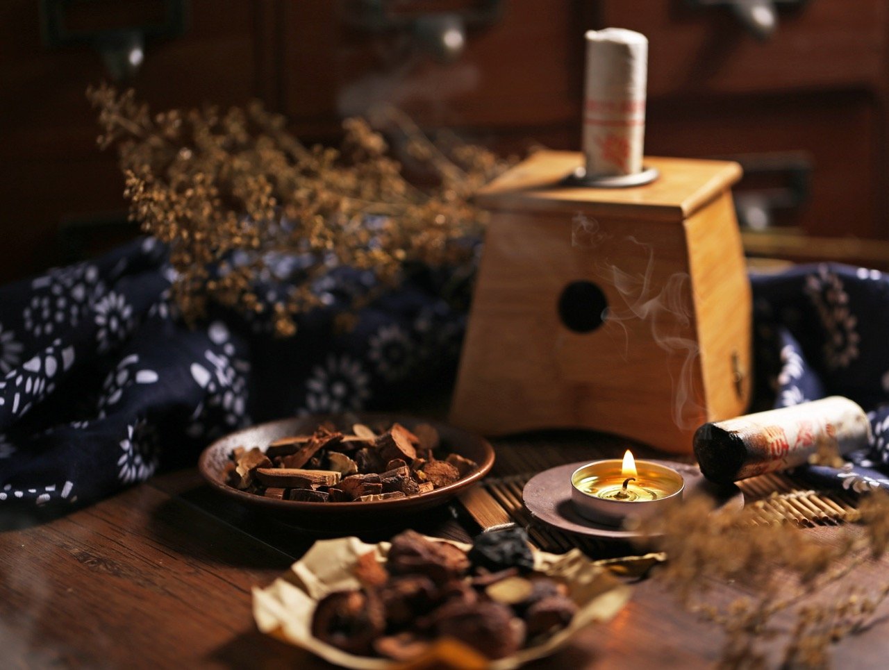 A wooden spice grinder with a lit candle beside it, surrounded by dried herbs, spices, and a bouquet of dried flowers on a dark, floral-patterned cloth.