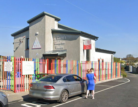A two-story childcare center building with a colorful picket fence in front. Two people are standing beside a parked gray car in the parking lot, with a woman in a blue dress and a child in a white and blue outfit. The building has signs that indicate it is a childcare center, and the sky is clear and blue.