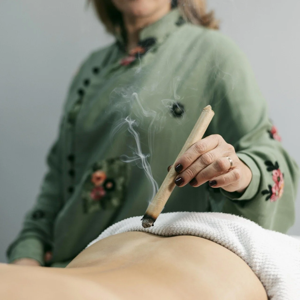 A person in a green jacket with floral embroidery performs a cupping therapy session, using a heated glass cup to create suction on a person's back.