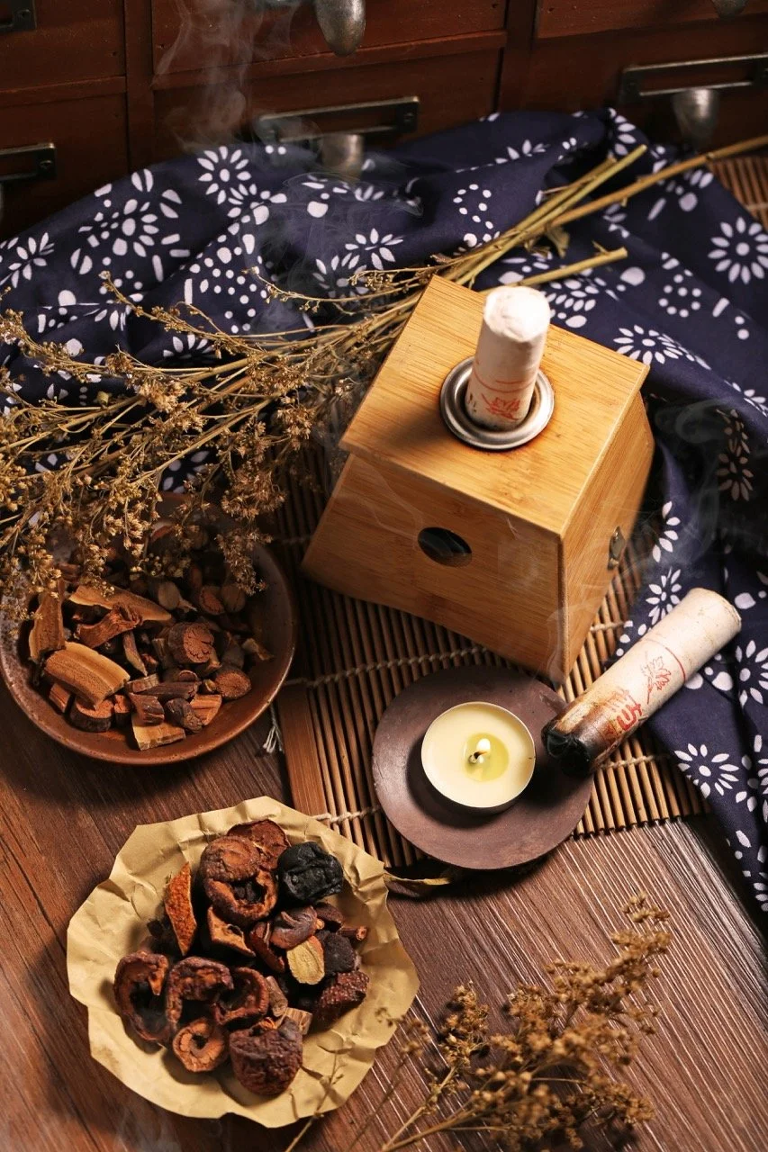 A wooden incense burner with smoke rising from it, surrounded by a bowl of dried herbs and spices, a candle, dried flowers, and a plate of dried fruit or herbs, all arranged on a bamboo mat with a blue and white patterned cloth.