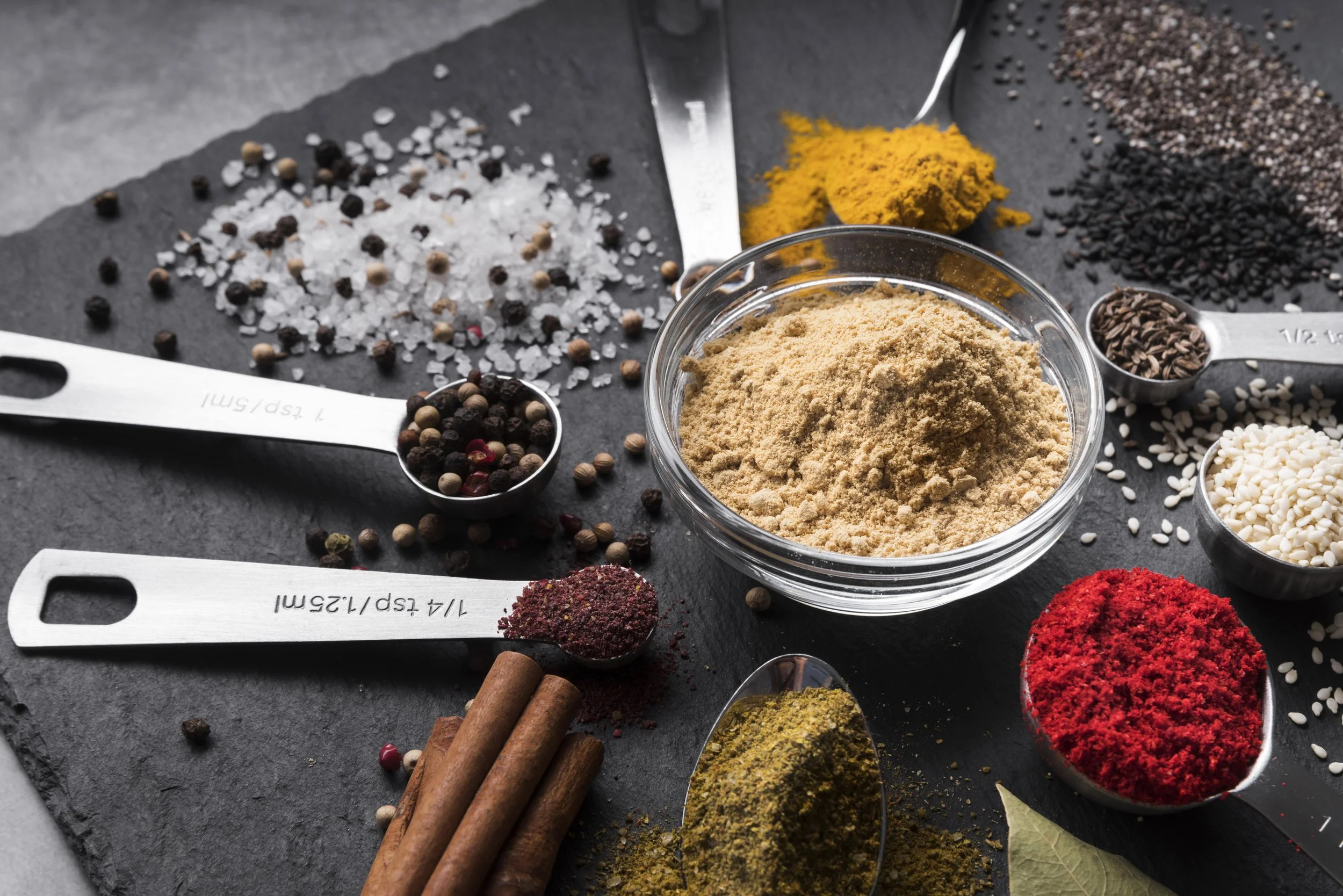 Assorted spices on a dark slate surface, including ground spice in a glass bowl, whole peppercorns, turmeric powder, black and white sesame seeds, red spice, and cinnamon sticks.
