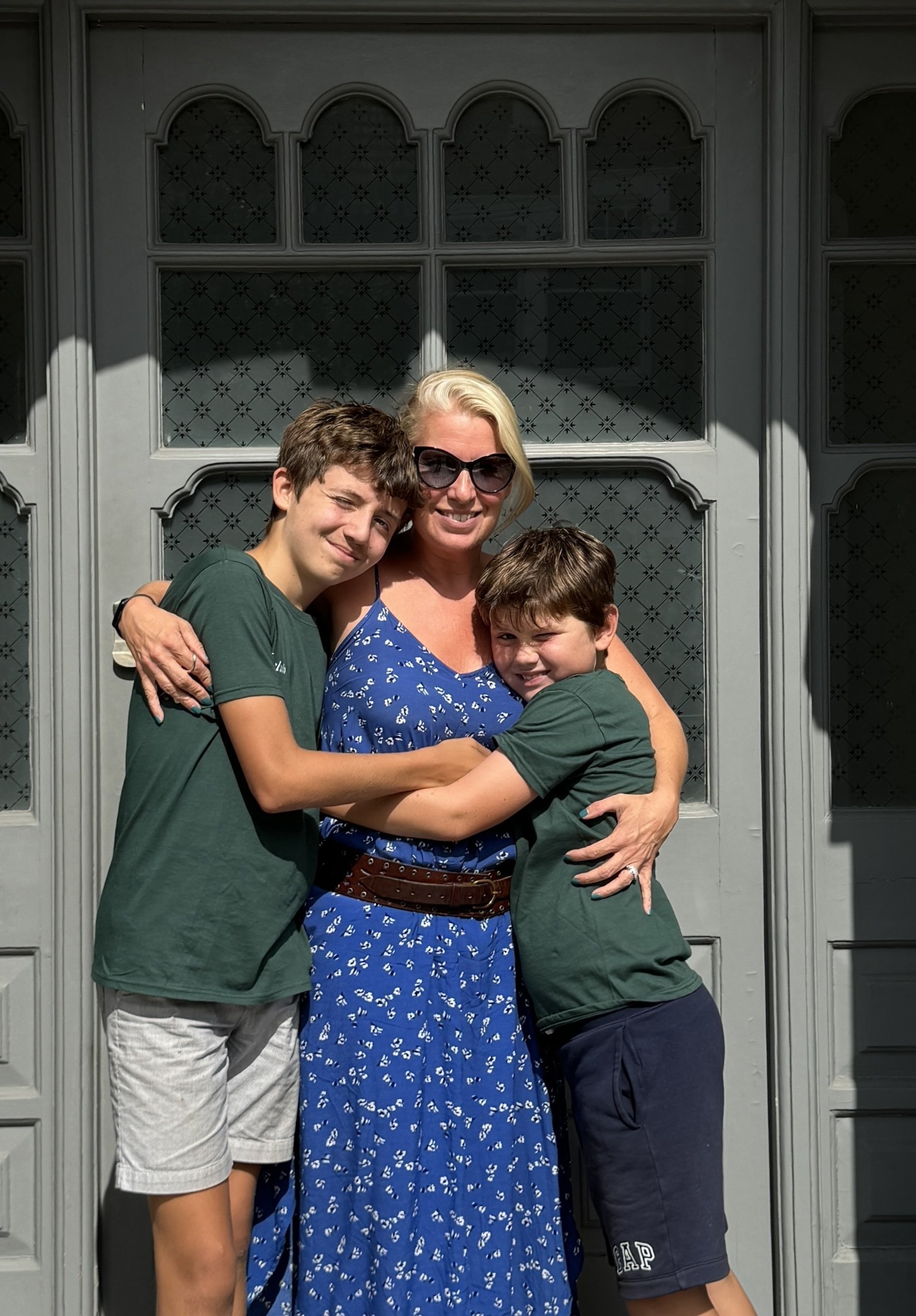 A woman with blonde hair, sunglasses, and a blue dress with white patterns is hugging two boys in front of a large gray door with decorative glass windows. The boys, wearing green shirts and dark shorts, are hugging her, one with his arm around her waist and the other with his arms around her neck. The sunlight casts shadows on the door and all three are smiling.