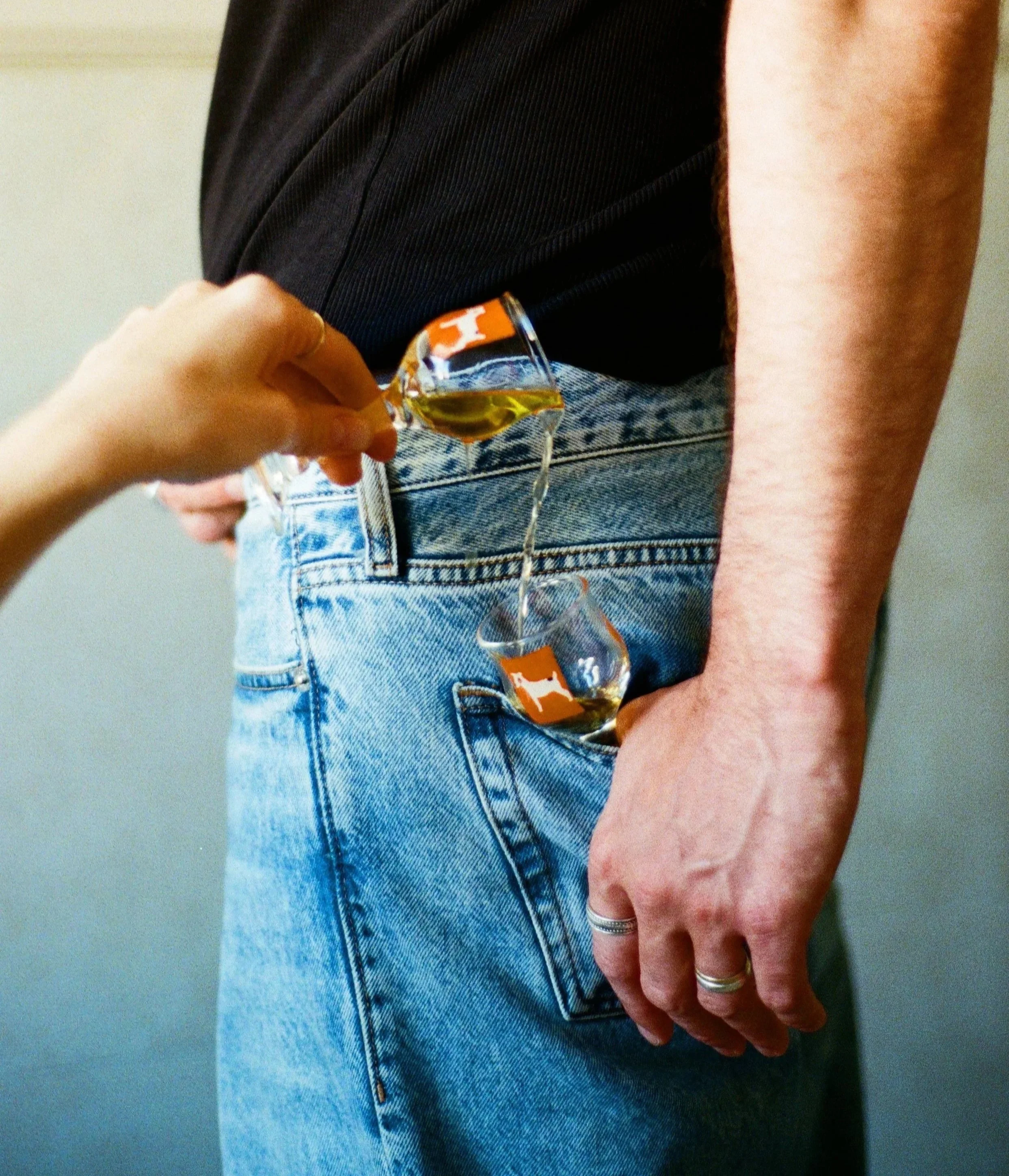 Close-up of a person's hand resting on their denim-clad hip, wearing multiple rings, with part of a black shirt visible, and holding a glass of yellow drink with ice.