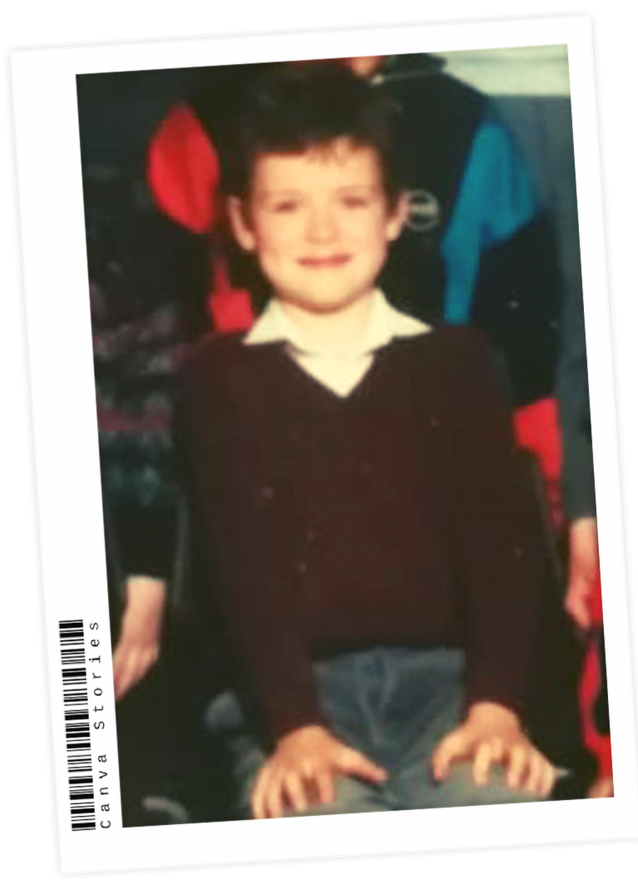 A young boy with short dark hair, wearing a white collared shirt, a dark sweater, and jeans, smiling while sitting at a table.