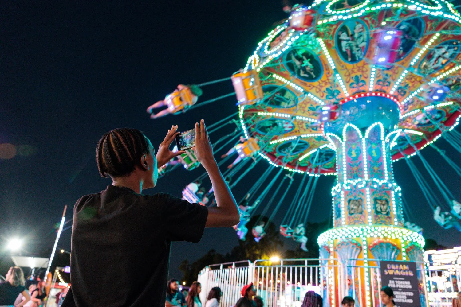 A person taking a photo of a brightly lit amusement park ride at night, with the ride spinning and colorful lights visible in the background.