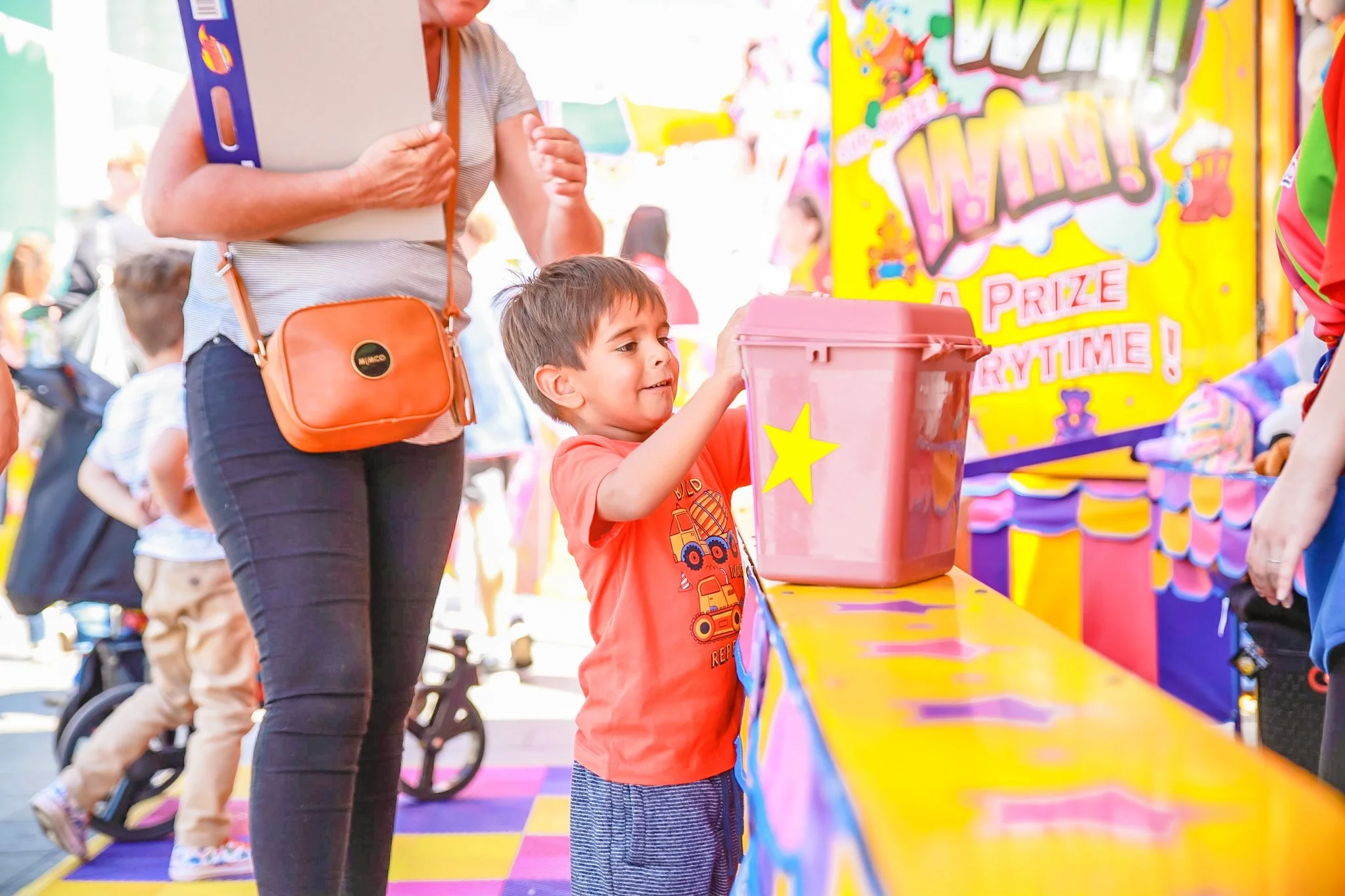 A young boy at a carnival game booth, holding a pink plastic container with a yellow star sticker. Behind him is a brightly colored, fun banner and other children and adults at the event.