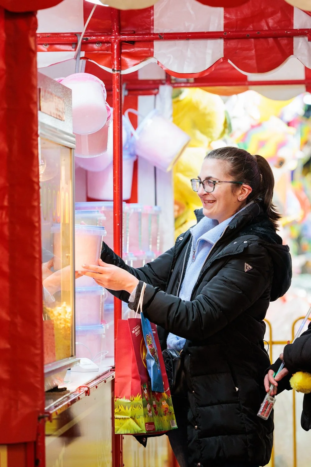 A woman with glasses smiling and shopping at a fair or market stall, holding a container of pink candy or popcorn. She is wearing a black jacket and a light blue hoodie, with a colorful shopping bag hanging from her arm. The stall features red and white striped fabric and various brightly colored items in the background.