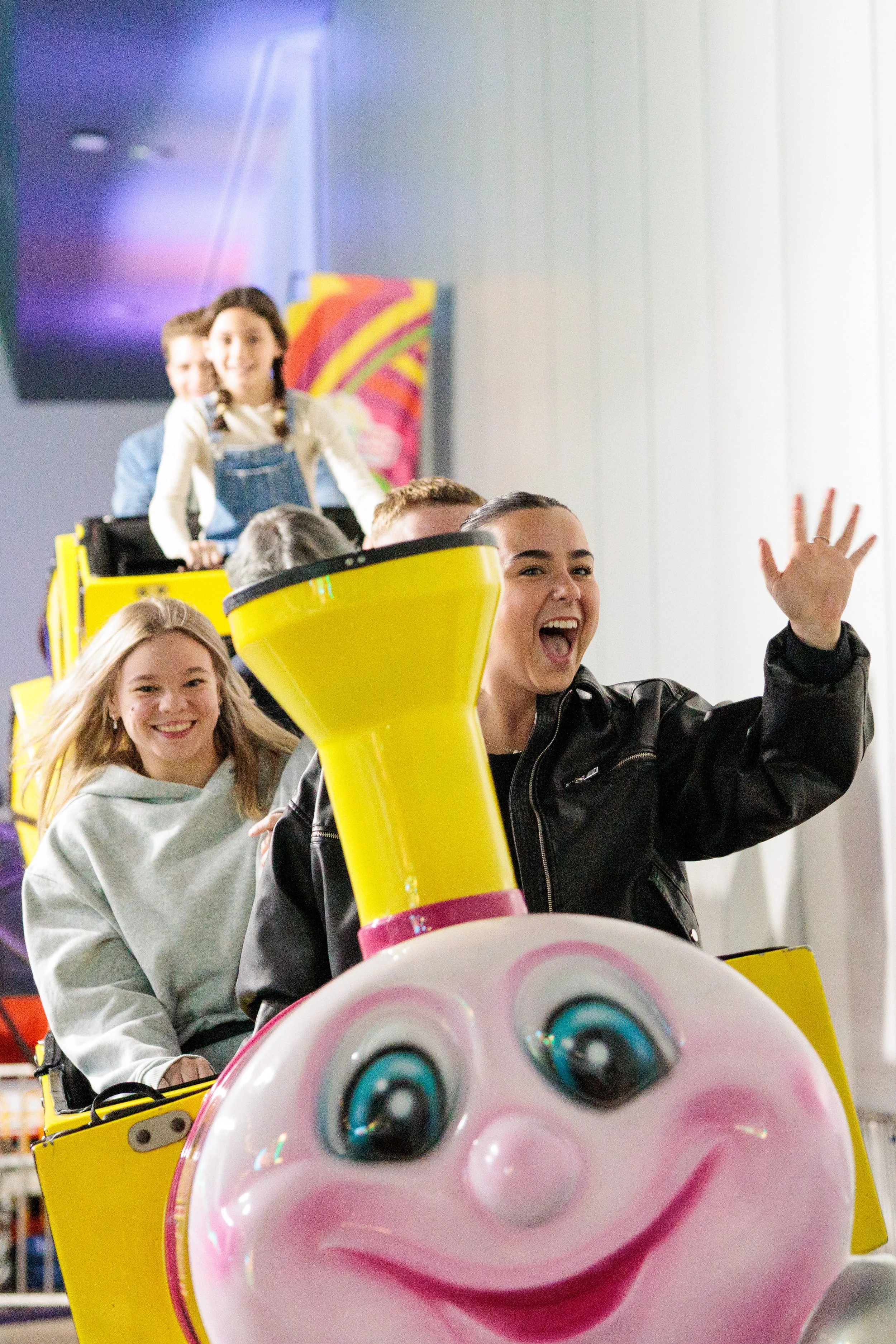 People riding a colorful amusement park ride with a pink cartoon pig face at the front, smiling and waving, in an indoor setting.