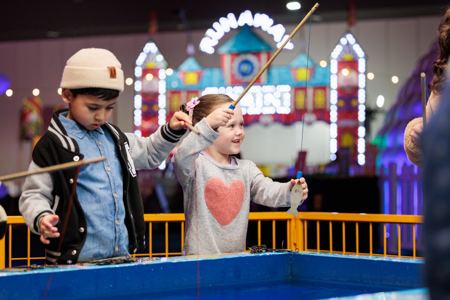Two children, a boy and a girl, fishing at an arcade game booth with a carnival theme, colorful lights, and a bright background.