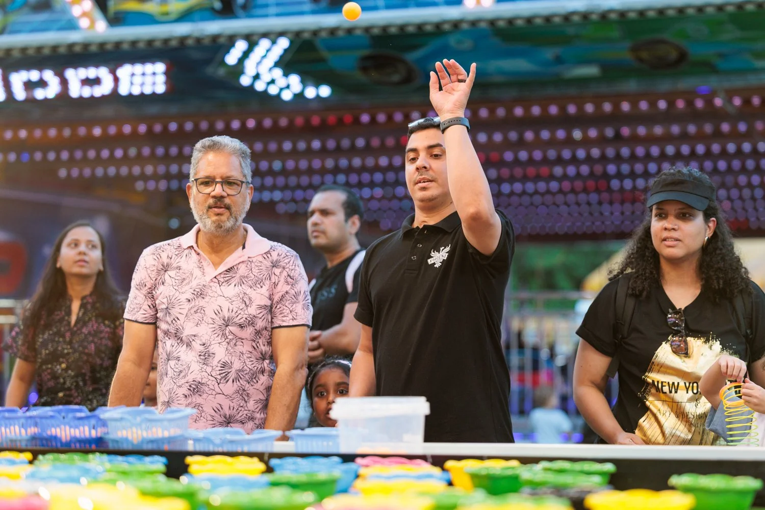 Group of people at a carnival or fair, with colorful lights and games in the background. Among them are a man in a pink floral shirt, a young man raising his hand, and a woman wearing sunglasses and a black cap.