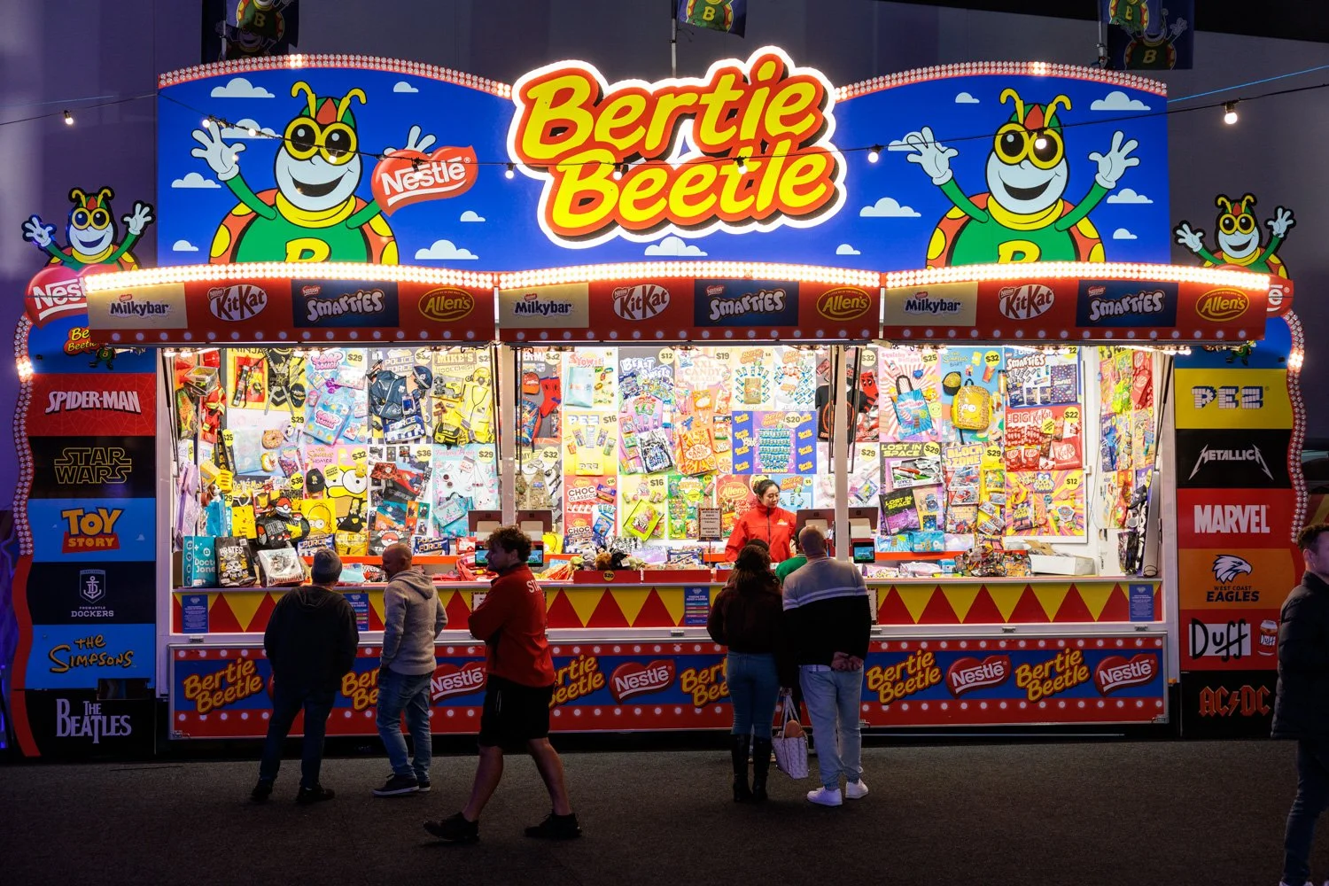 A colorful carnival game booth featuring Bertie Beetle branding, with plush toys, candy, and novelty items displayed inside. The booth is decorated with large animated Bertie Beetle cartoon characters, bright lights, and logos of popular brands such as Nestlé, Kit Kat, Smarties, and Allen's. Several people are gathered around, some paying for prizes or browsing the merchandise.