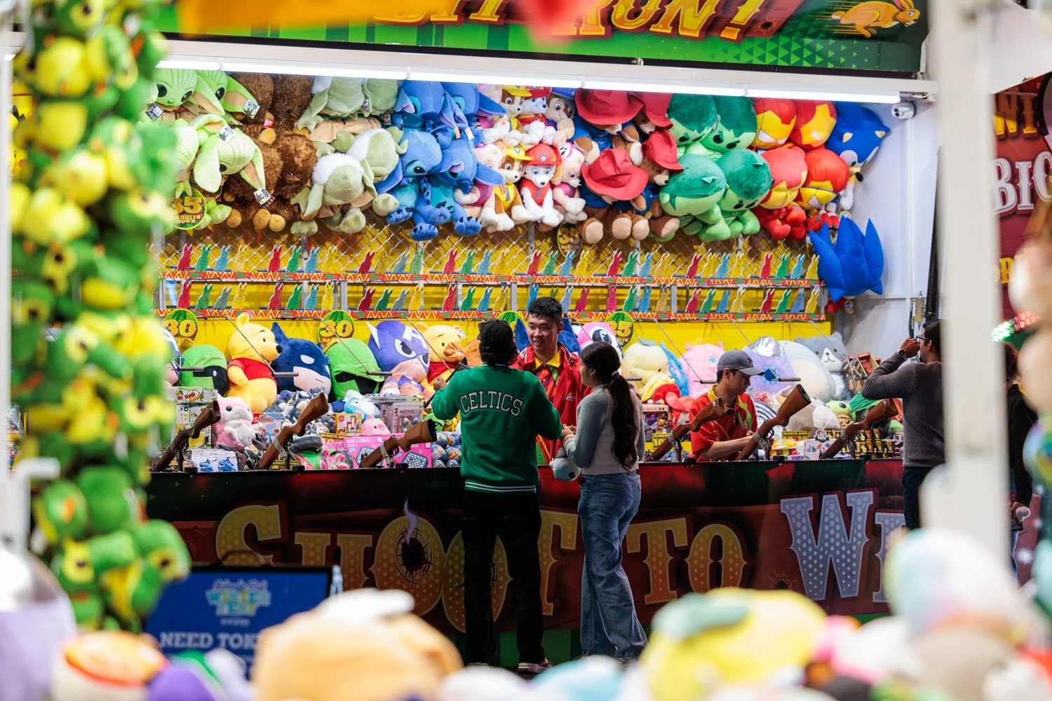 People playing carnival ring toss game with plush toys as prizes, large stuffed animals displayed behind the booth, colorful decorations and banners, and game attendants assisting players.