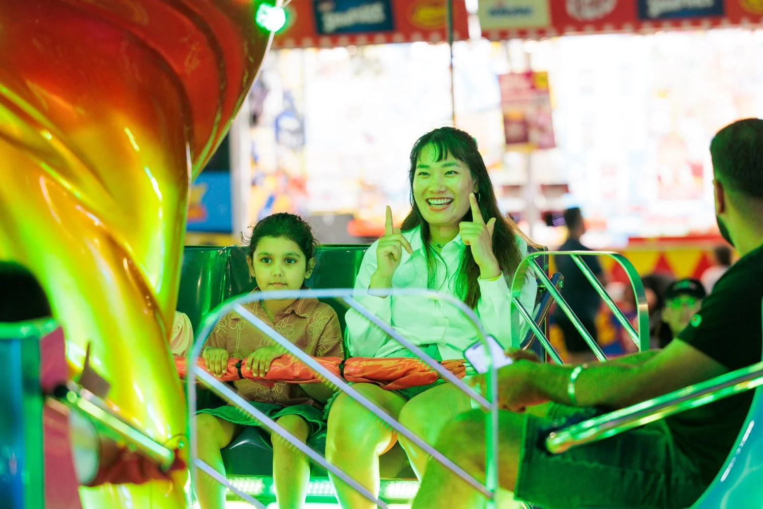 A woman and a young girl sit on an amusement park ride with colorful neon lights. The woman is smiling and pointing with her fingers, while the girl looks serious. The background shows a bright, festive amusement park scene.