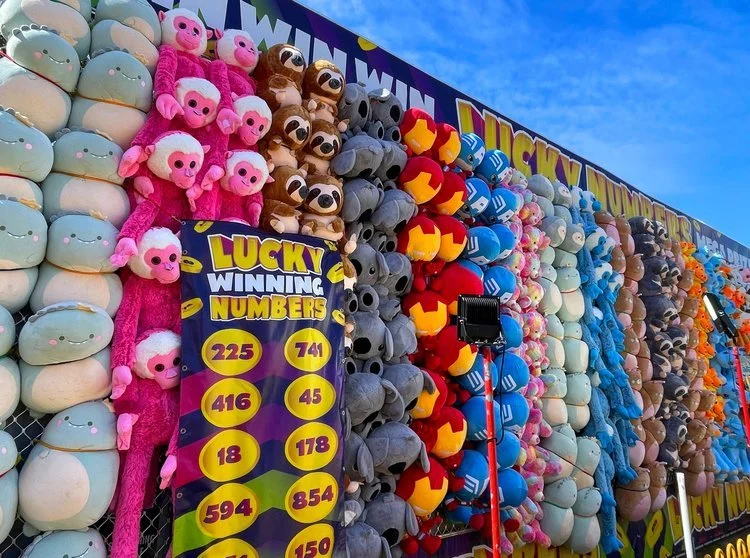 Colorful stuffed animal prizes at a carnival game, including pink monkeys, koalas, Spiderman, Blue Power Rangers, Captain America, and other plush toys, with a sign displaying winning numbers.