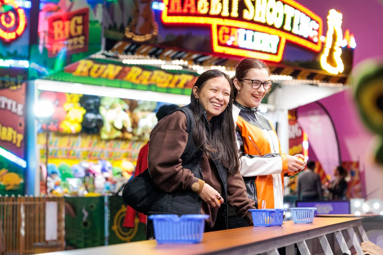 Two women smiling and enjoying themselves at an amusement park or fairground. The background features bright neon signs and colorful carnival game booths.