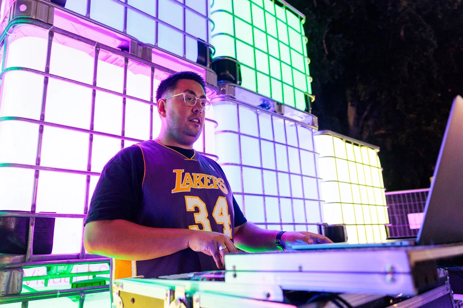 A man wearing a Los Angeles Lakers basketball jersey and glasses is DJing at a vibrant outdoor event with colorful illuminated grid structures in the background.