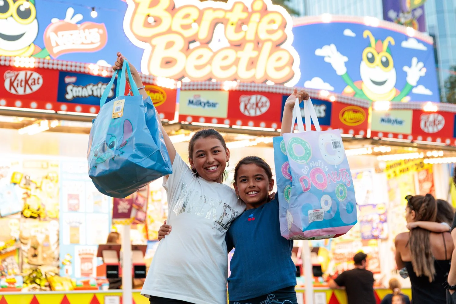 Two young girls smiling and holding plush bags at a carnival with a colorful sign that reads "Bertie Beetle" in the background.