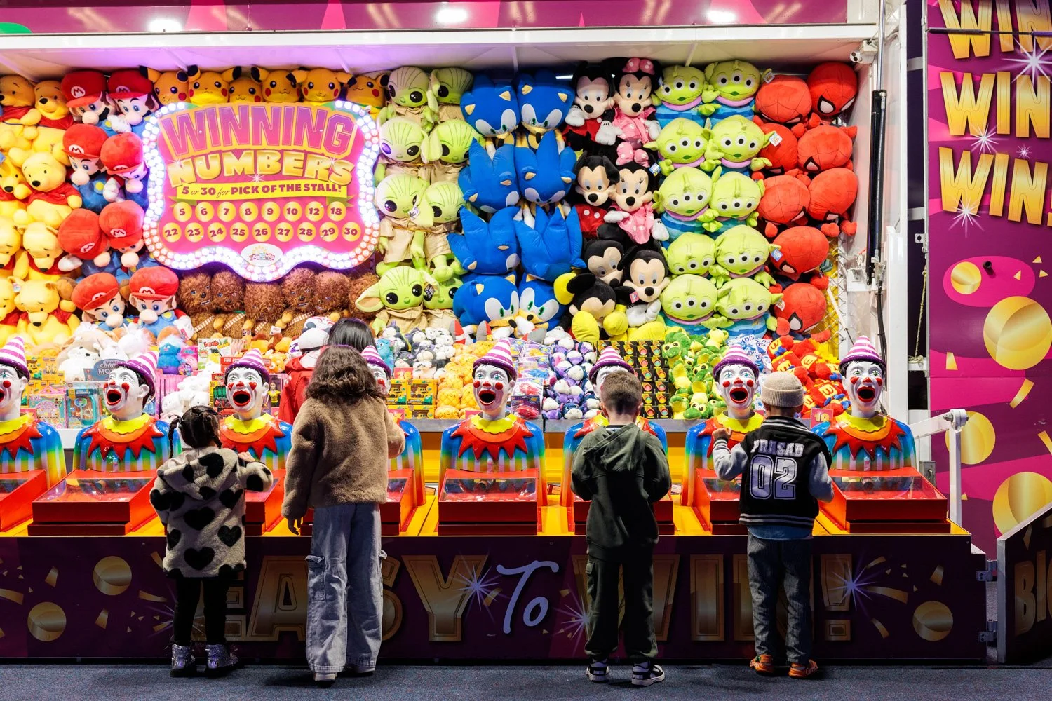 Children playing a carnival game with clown heads on the prize table, surrounded by plush toys including Mario, Pikachu, Sonic, and Disney characters, under bright lights with a colorful sign reading 'Winning Numbers' and a purple and pink background.