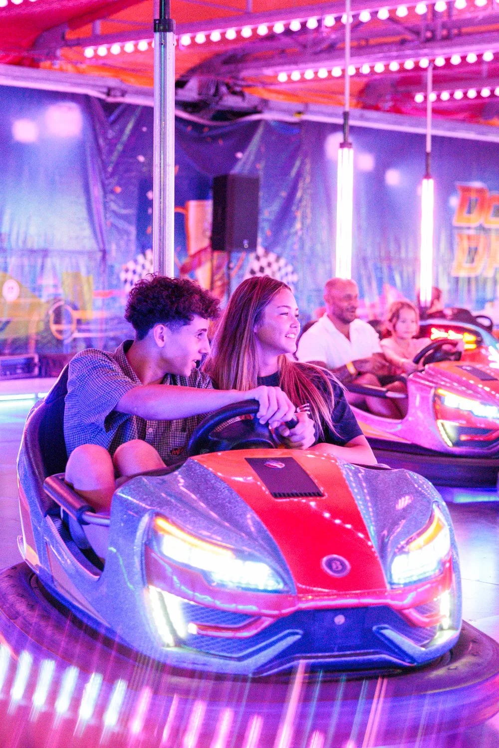Young people riding a colorful bumper car at an indoor amusement park with bright neon lights.