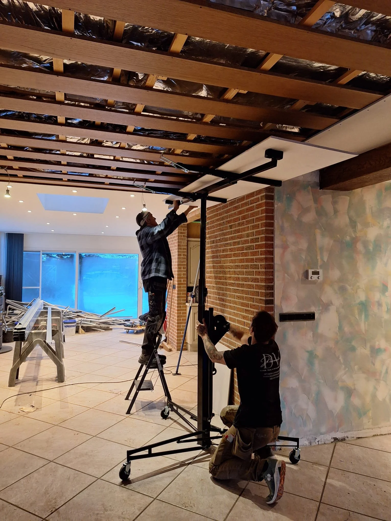 Mannen hangen een plafond tussen houten balken op, met behulp van een ondersteuning en een ladder. Een andere man zit op de grond, houdt een apparaat of gereedschap vast en kijkt naar de mannen die het plafond installeren. De locatie is een binnenruimte met een gemetselde muur, een kleurrijk geschilderde muur en grote ramen.