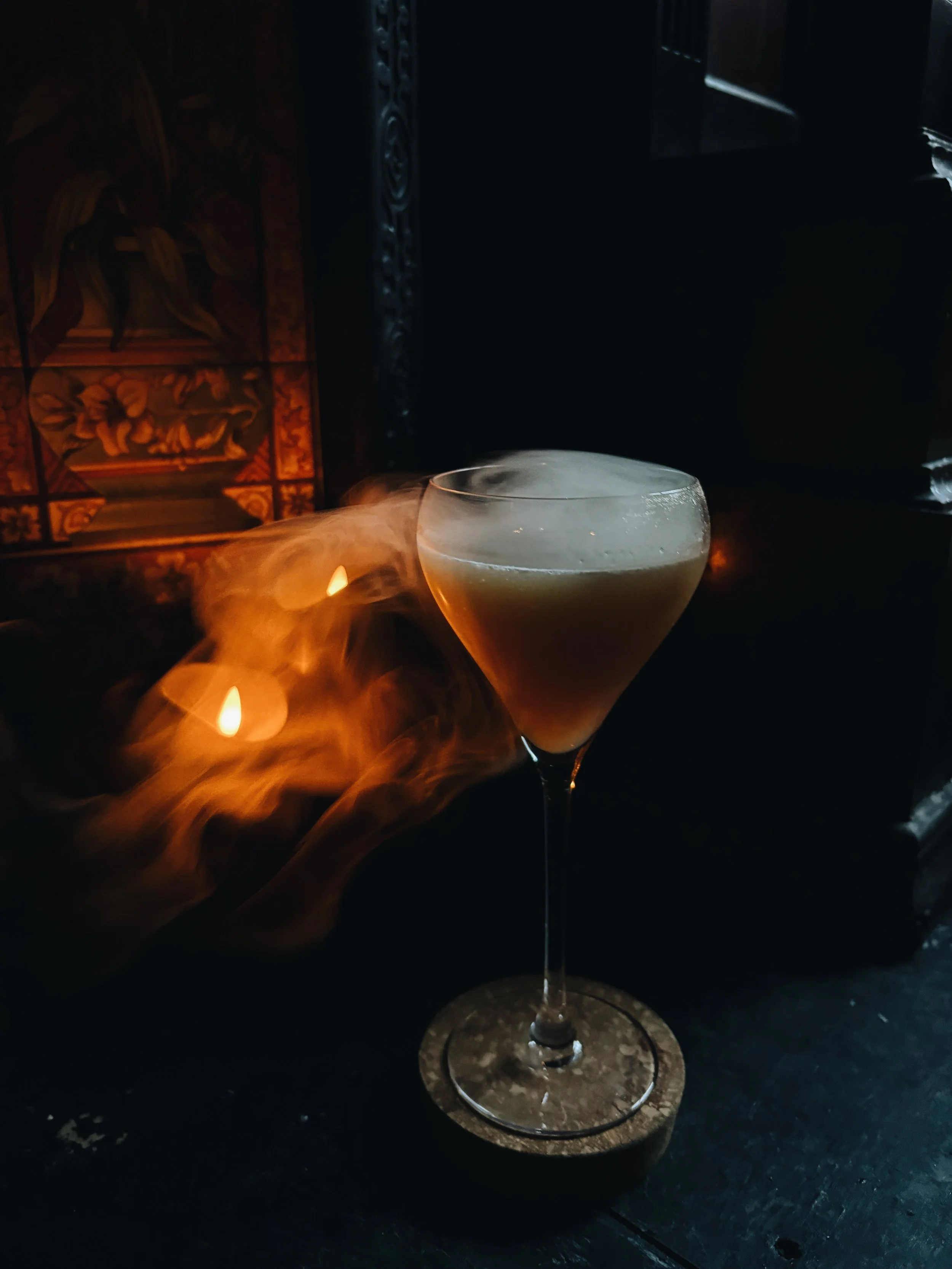 A cocktail glass with a stem, filled with a drink, is placed on a dark surface. There is steam rising from the drink. In the background, there are candles and a decorative element, which seems to be traditional or artistically carved. The lighting is