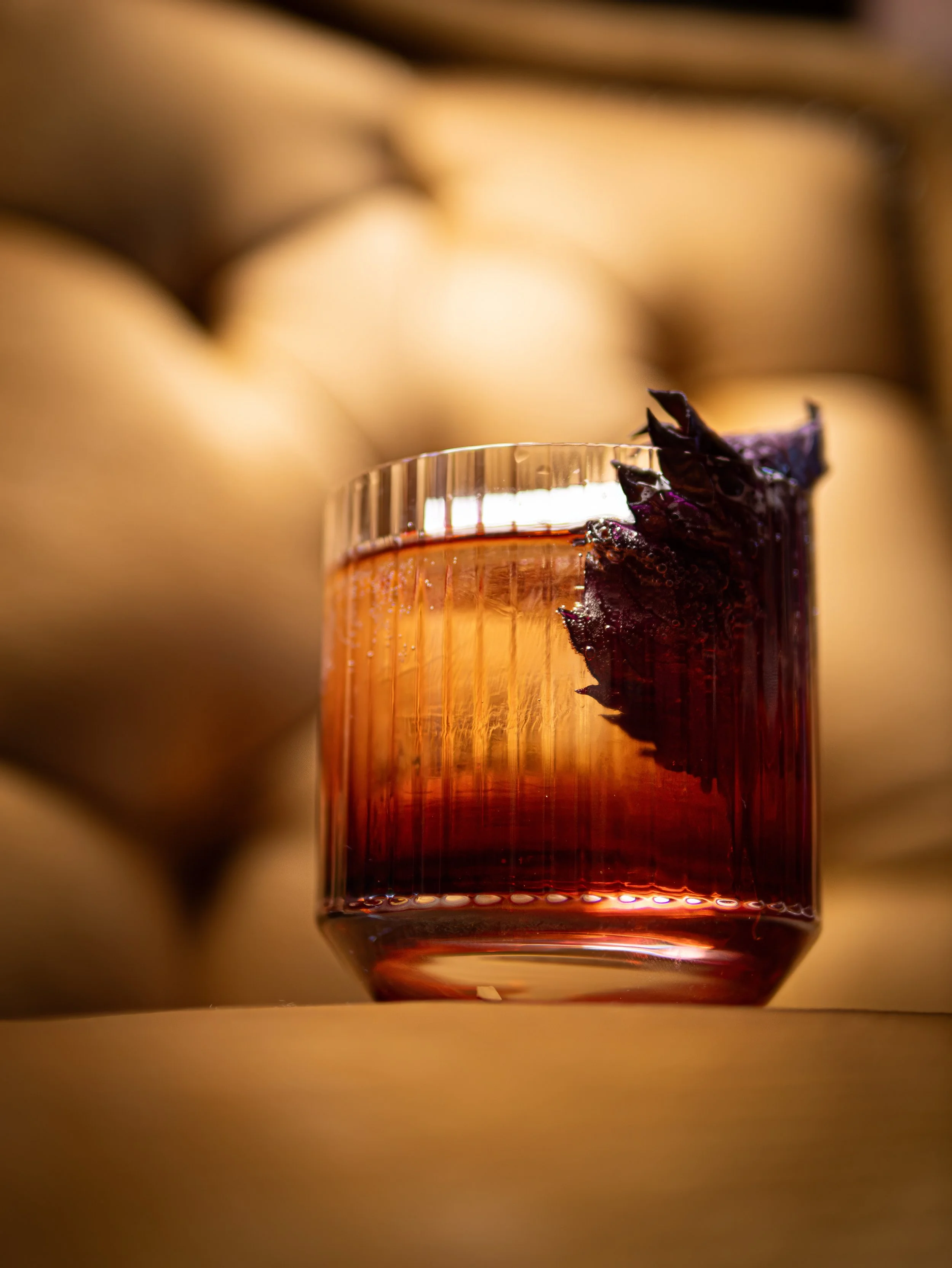 Close-up of a glass of dark-colored liquid, possibly wine or juice, with a piece of purple or black fruit garnishing the rim. Out-of-focus background showing blurred yellow and brownish colors.