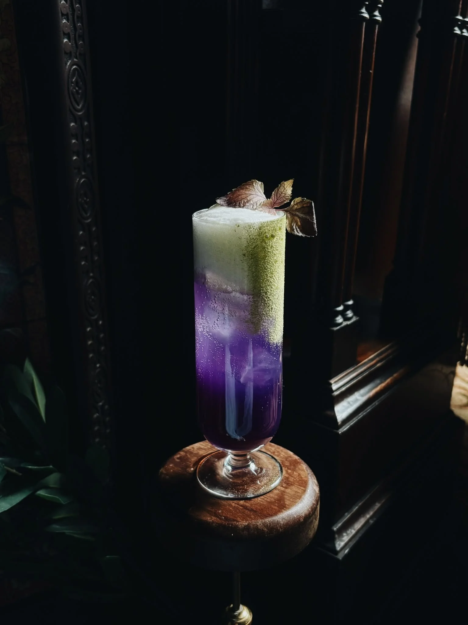 Colorful layered cocktail with purple, white, and green sections, garnished with a leaf, in a tall glass on a wooden surface.