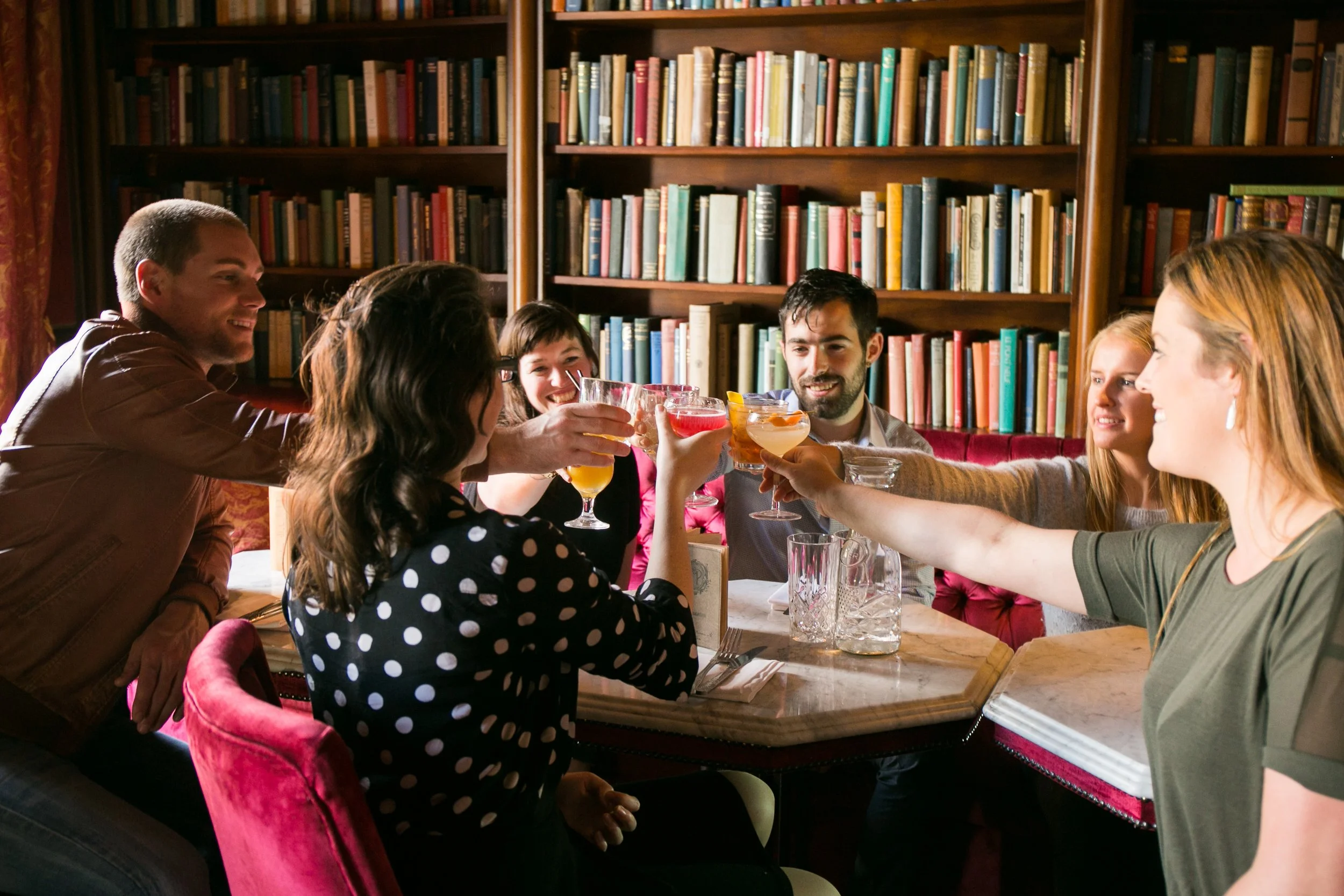 A group of friends sitting around a table in a cozy, book-filled room, raising their drinks in a celebratory toast and smiling at each other.