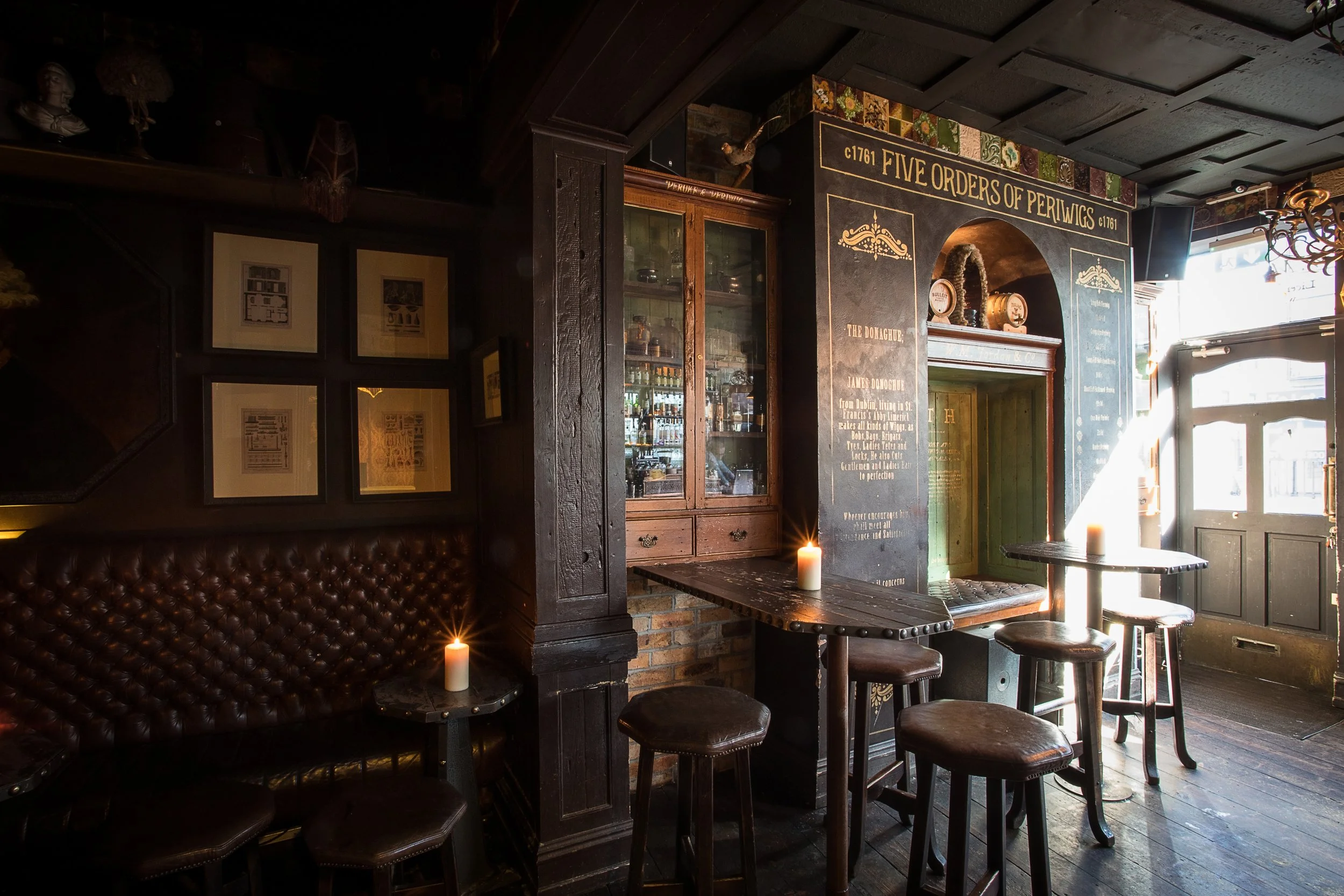 A cozy pub interior featuring a dark wood bar, framed pictures on the wall, and a table with chairs near a window with sunlight. Candlelight adds warmth to the scene.
