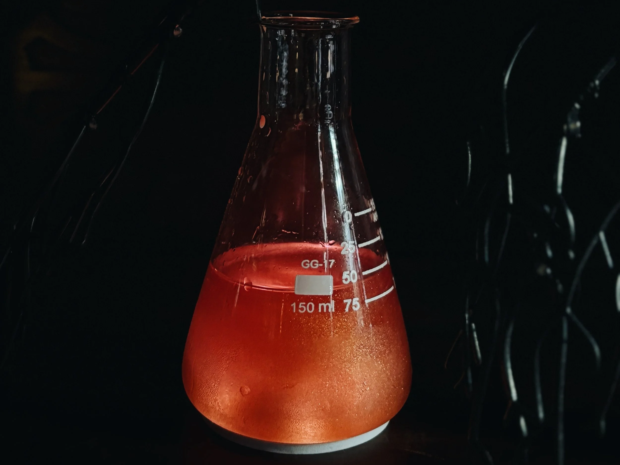 A laboratory glass beaker filled with red liquid, with measurement markings from 50 to 150 milliliters, set against a dark background.