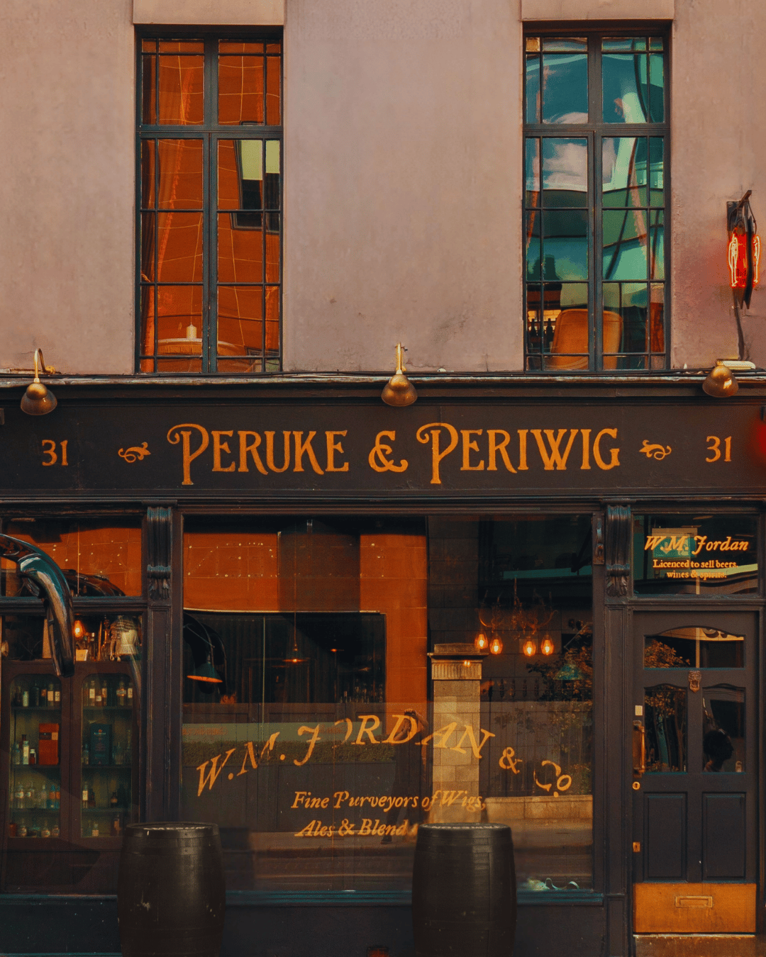 The storefront of Peruke & Periwig, a shop at 31, with black and gold signage, large windows reflecting nearby buildings, and barrels in front.