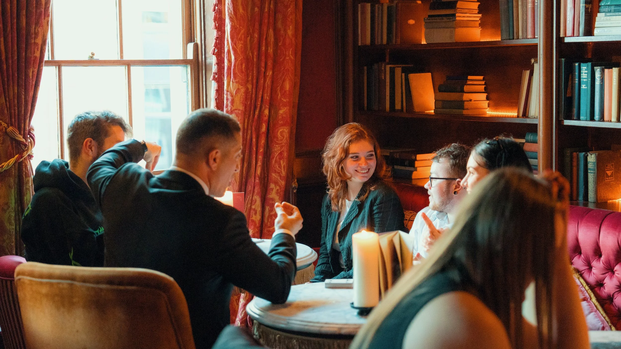 Group of people gathered around a table in a cozy, dimly lit room with shelves of books, enjoying conversation and drinks.