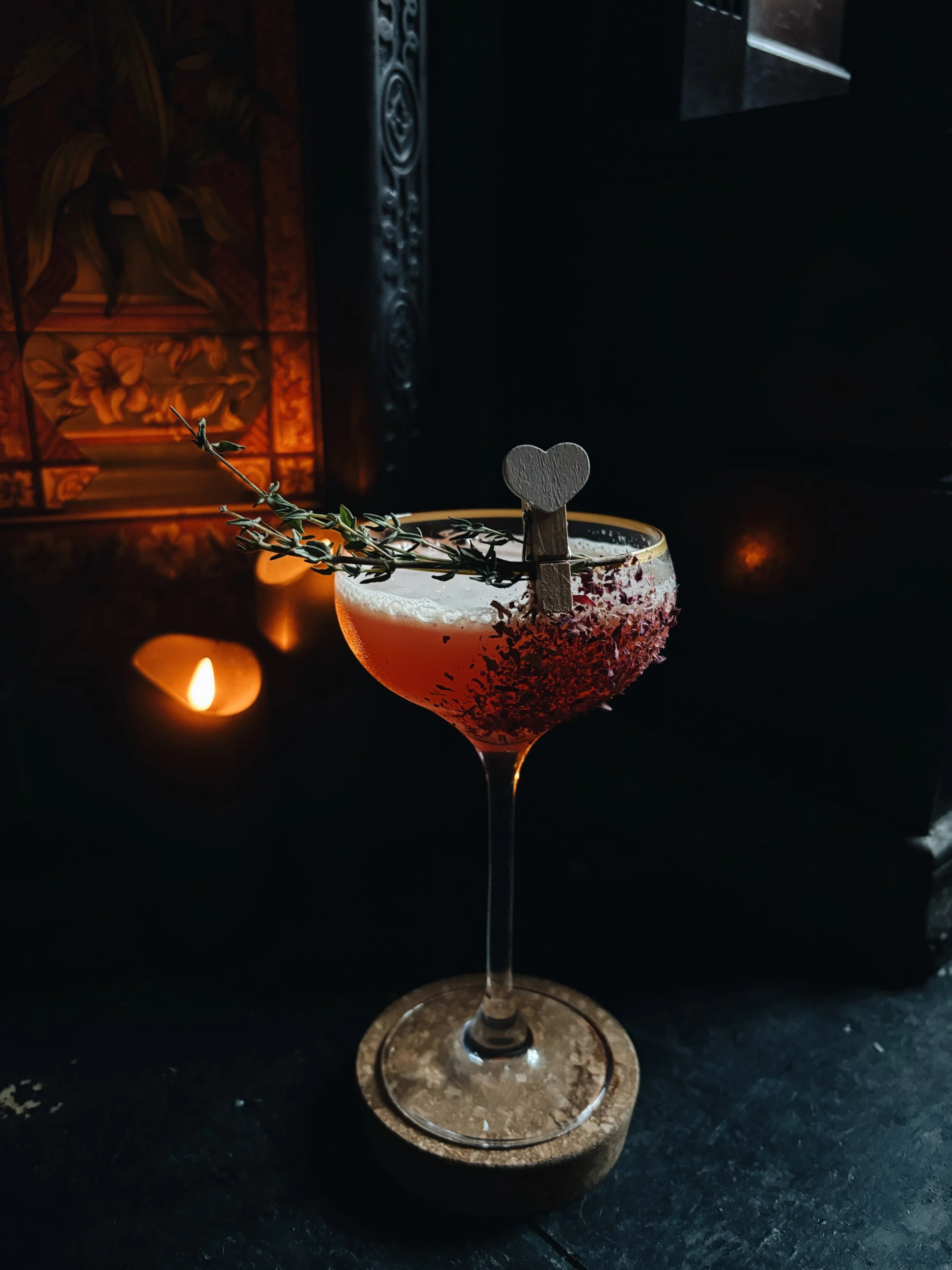 A cocktail in a stemmed glass with a salted rim, garnished with a sprig of thyme and a small wooden heart on top. The background features warm lighting and decorative elements.