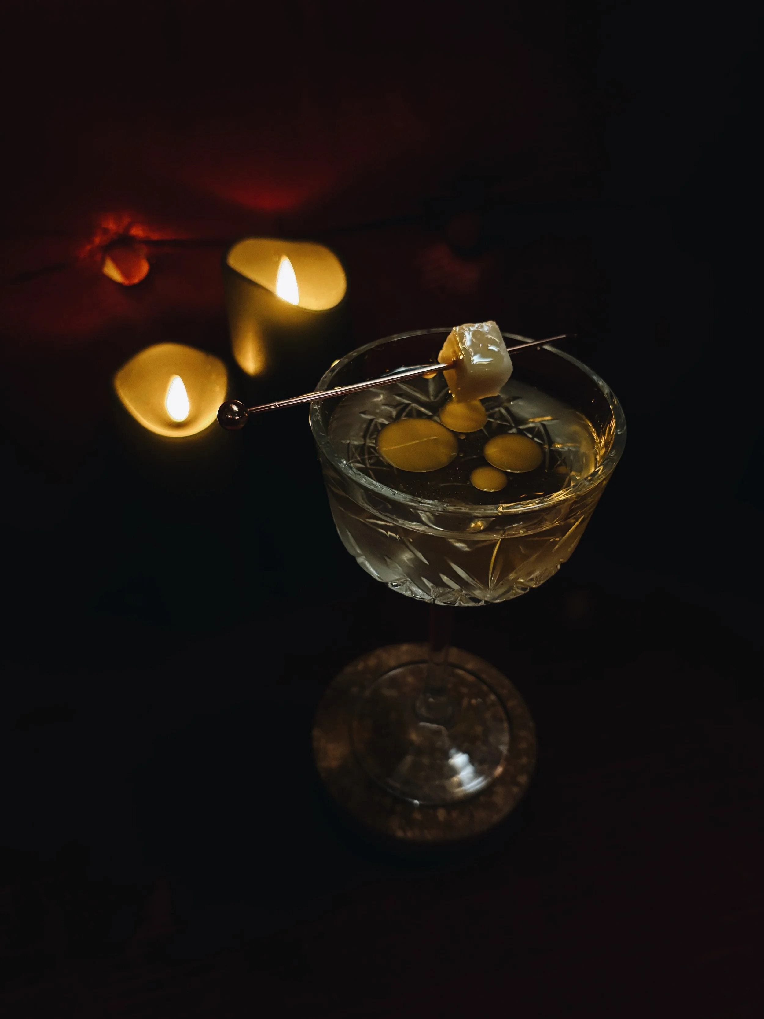 A glass filled with whiskey, with a cocktail pick holding a piece of cheese, in a dark setting illuminated by two candles in the background.