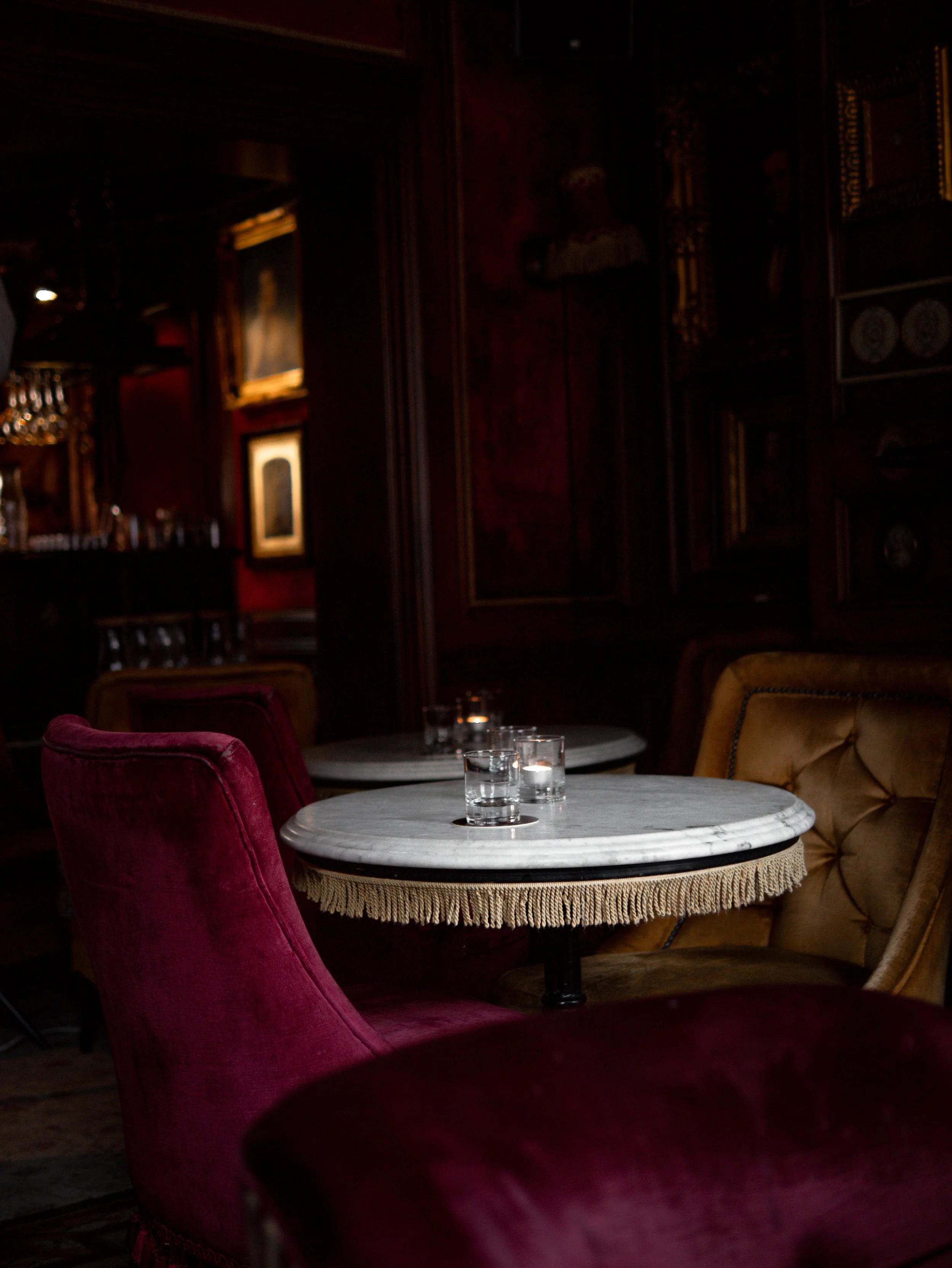 Dimly lit vintage-style restaurant or lounge with round marble-topped tables, plush velvet chairs in red and yellow, and dark wood paneling with framed artwork.