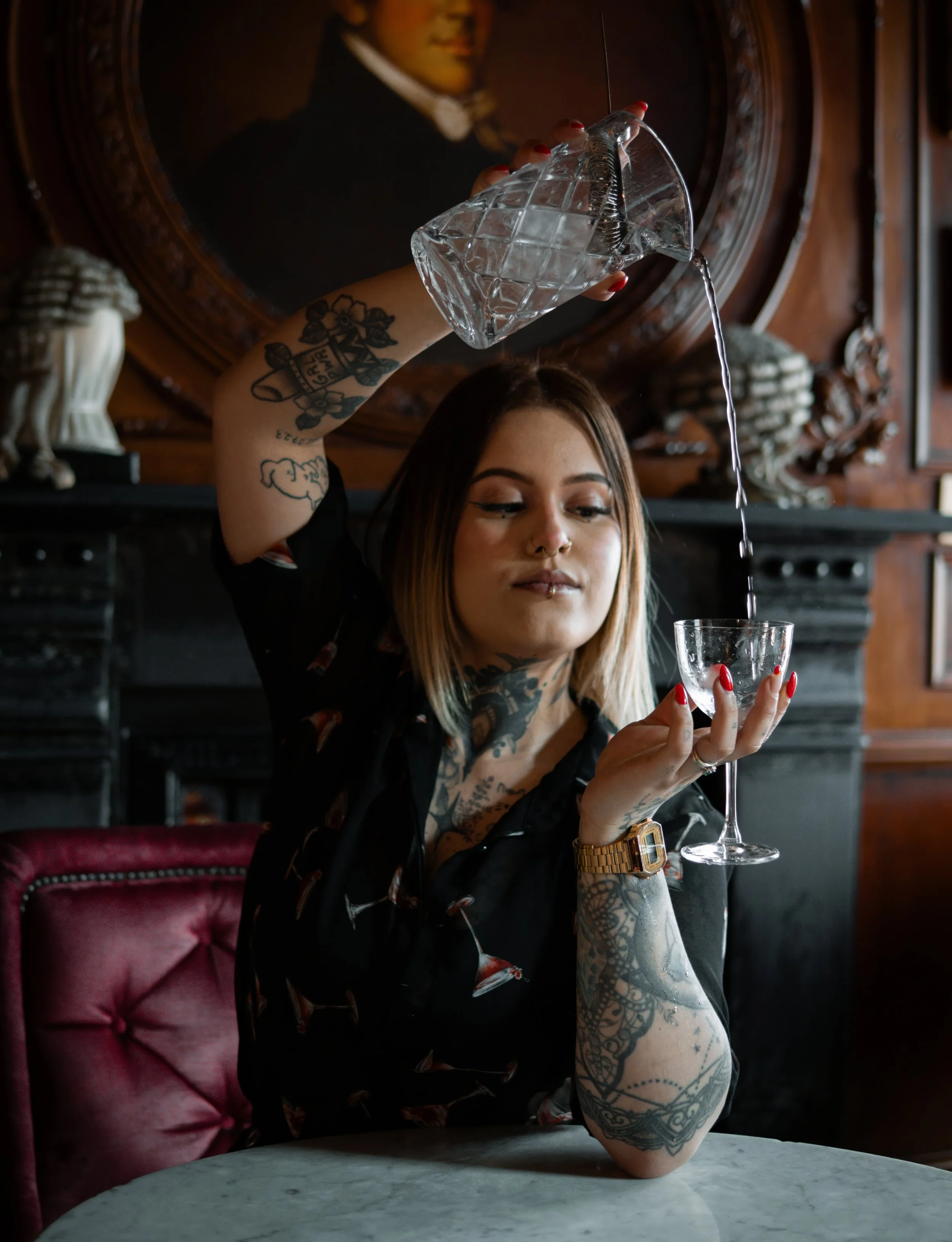 A woman with tattoos and a necklace sits at a marble table in a vintage-style room with a pink velvet chair, holding a cocktail.