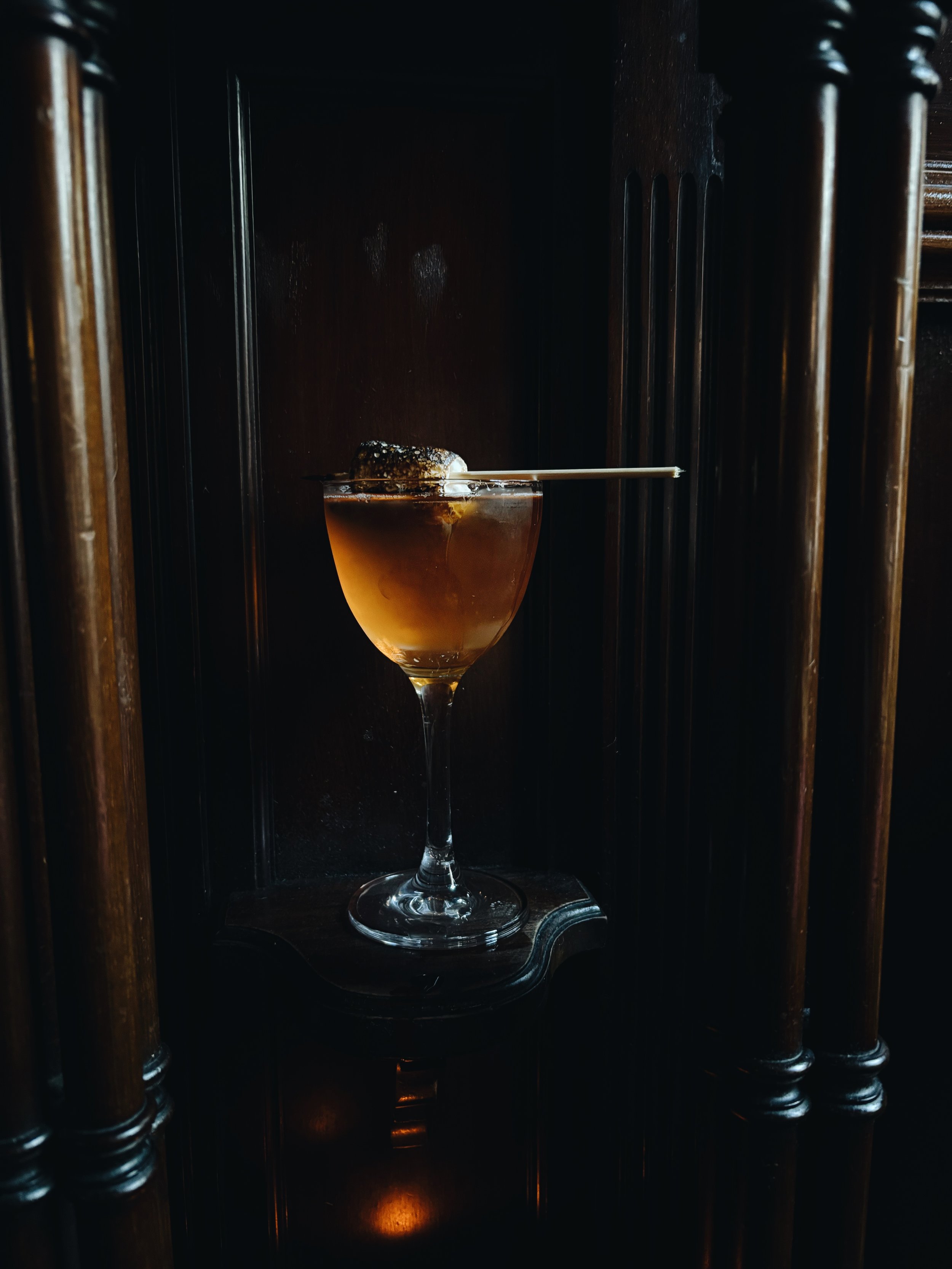 A flaming cocktail garnished with a charred marshmallow on a skewer, served in a coupe glass against a dark background.