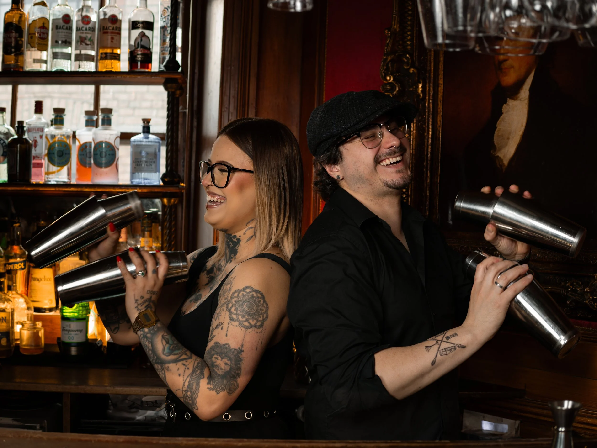 Two smiling bar staff, a woman with tattoos and glasses and a man with glasses and a cap, behind a bar with liquor bottles on shelves, using cocktail shakers.