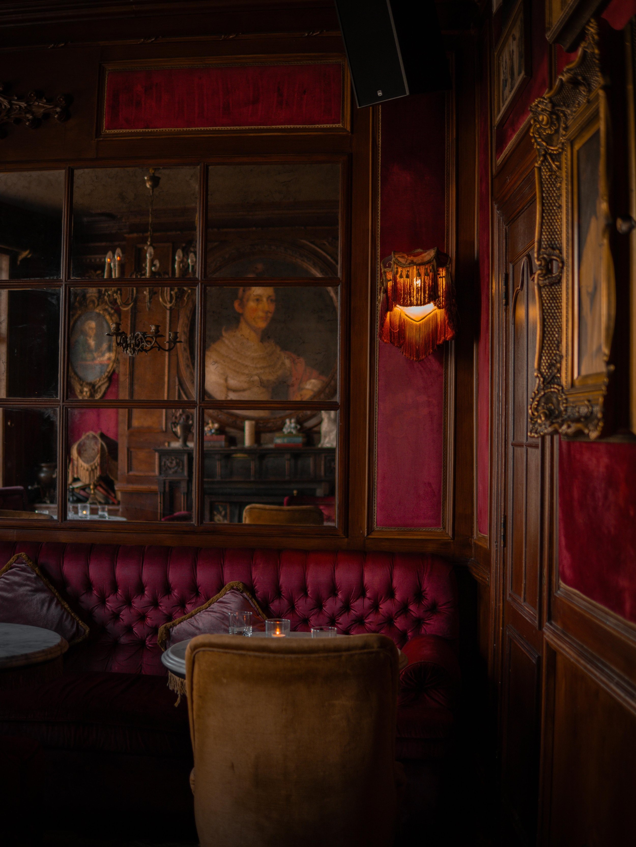 Interior of a luxurious room with red velvet padded walls, ornate wooden paneling, a velvet sofa with pillows, a small round table with glasses, and candle lighting. A reflection in a mirror shows a woman in period costume.