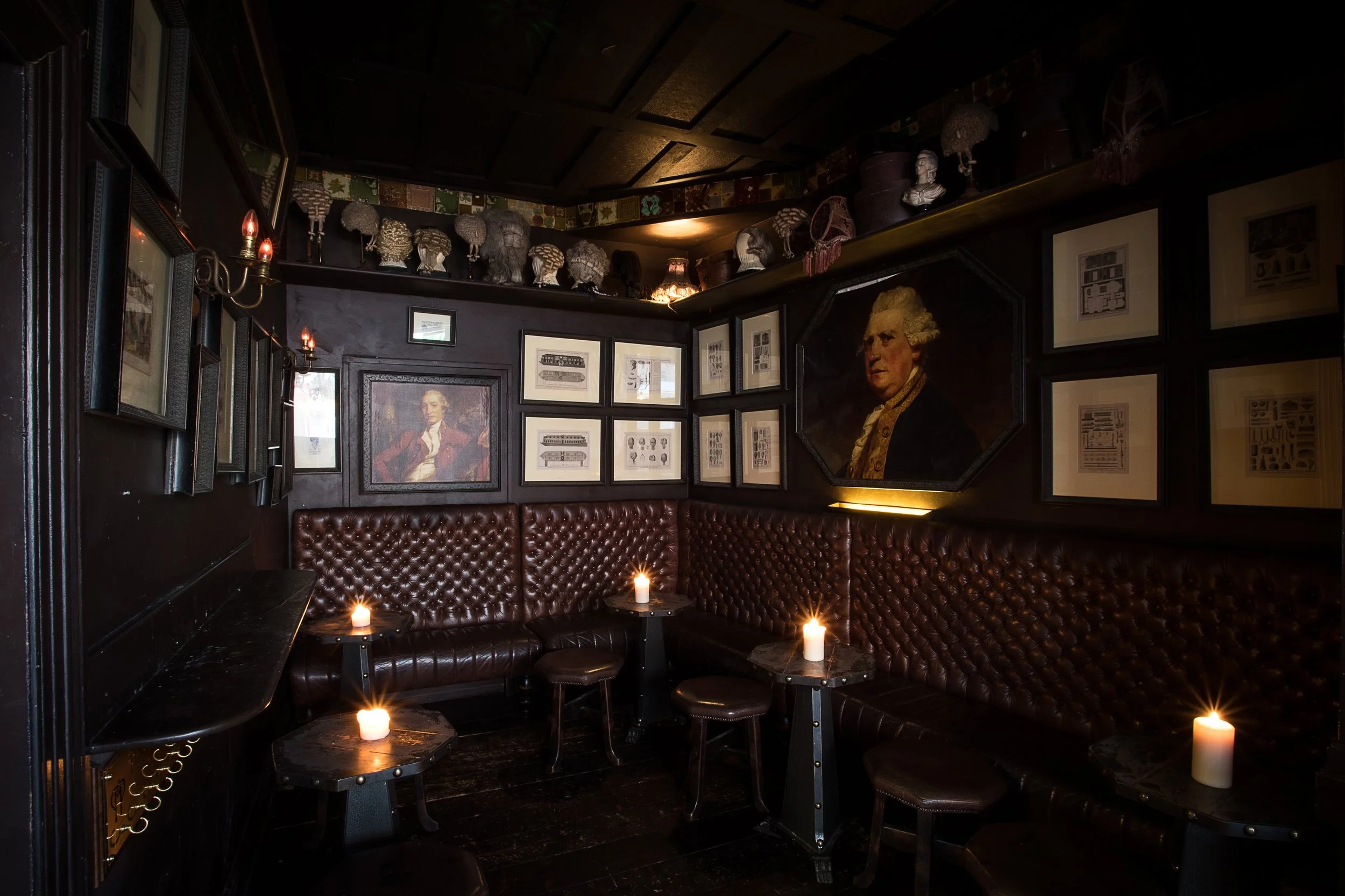 A cozy dimly lit room with dark wooden paneling and leather tufted seating, decorated with framed artwork and bust sculptures on the upper shelf. Small candles provide ambient lighting on tables.