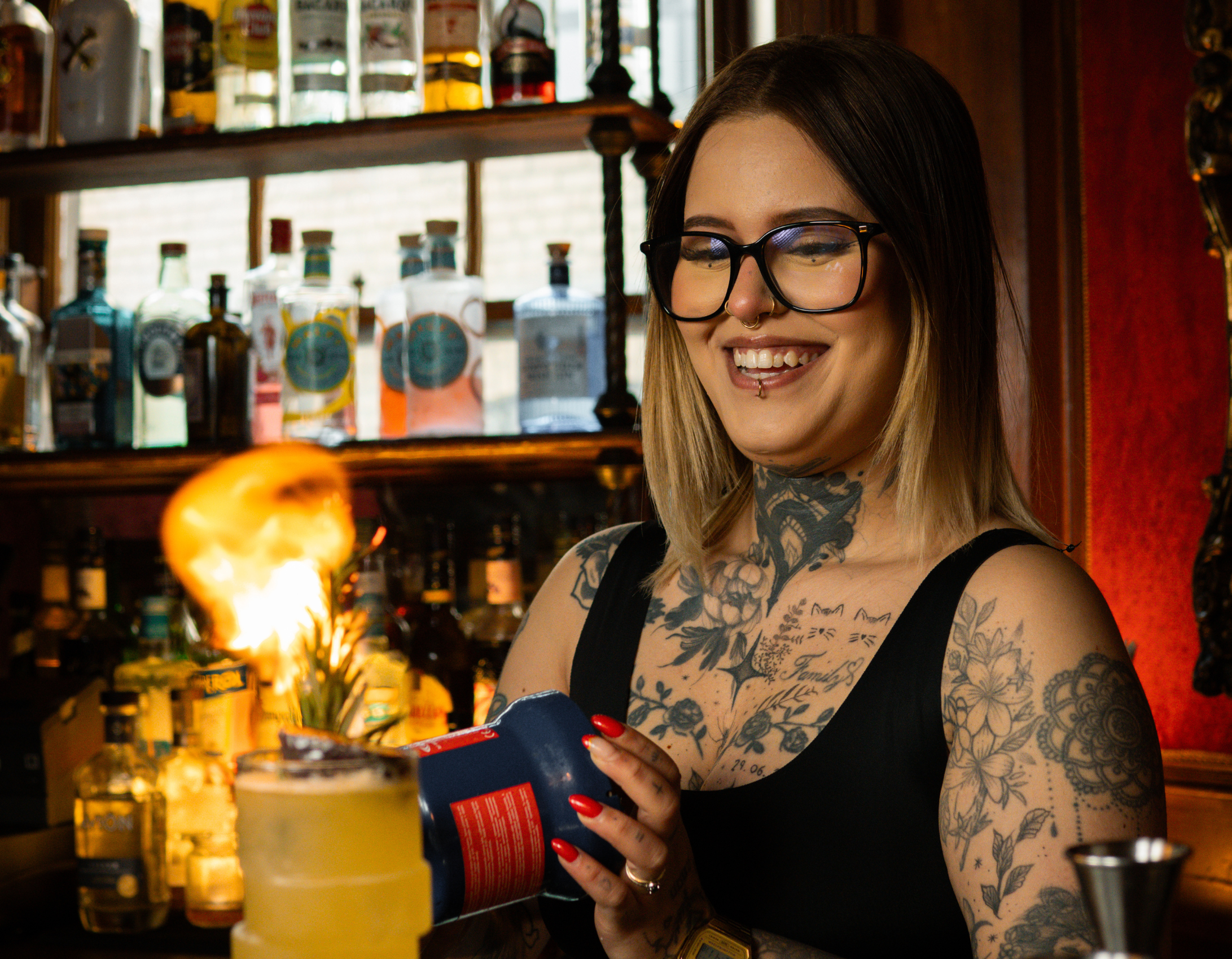A woman with glasses and tattoos smiling and holding a torch inside a bar, with bottles on shelves in the background.