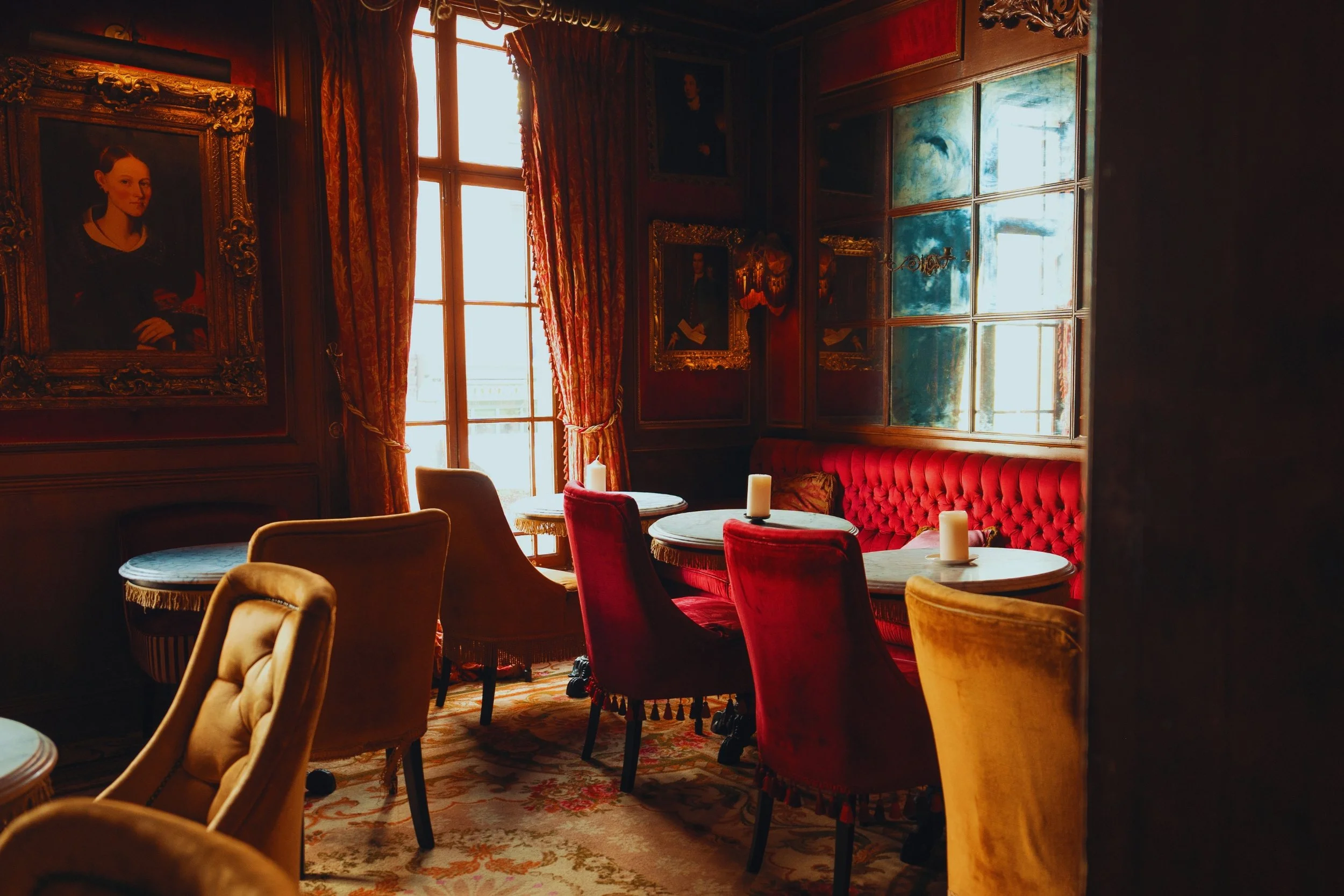 An elegant, vintage-style room with red velvet upholstery on a long bench, colorful upholstered chairs, and a table with candles. The room features ornate gold-framed portraits on dark wooden walls, large windows with patterned curtains, and a decorative carpet on the floor.