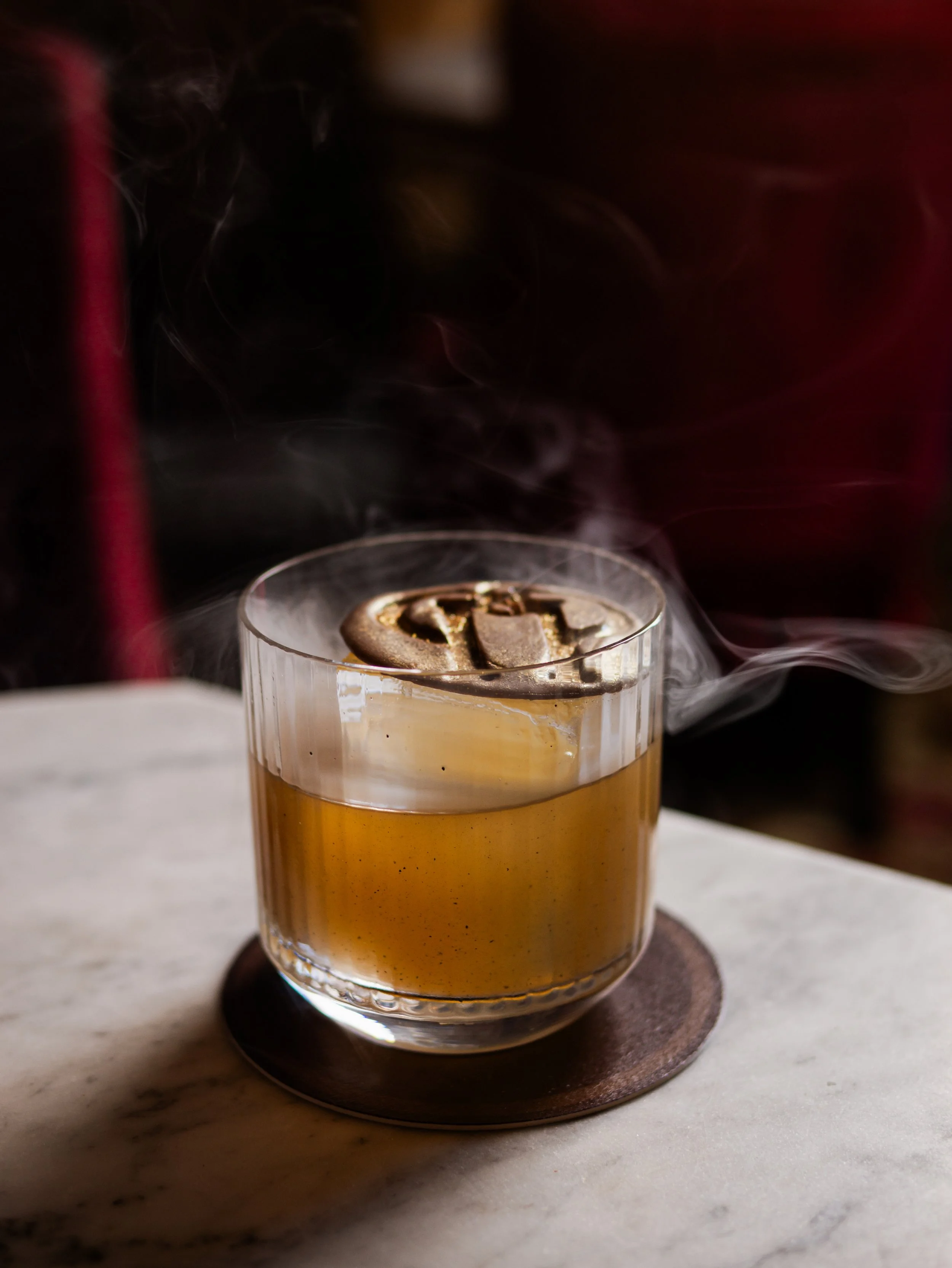Close-up of a cocktail with smoke rising on a marble surface, with a dark background.