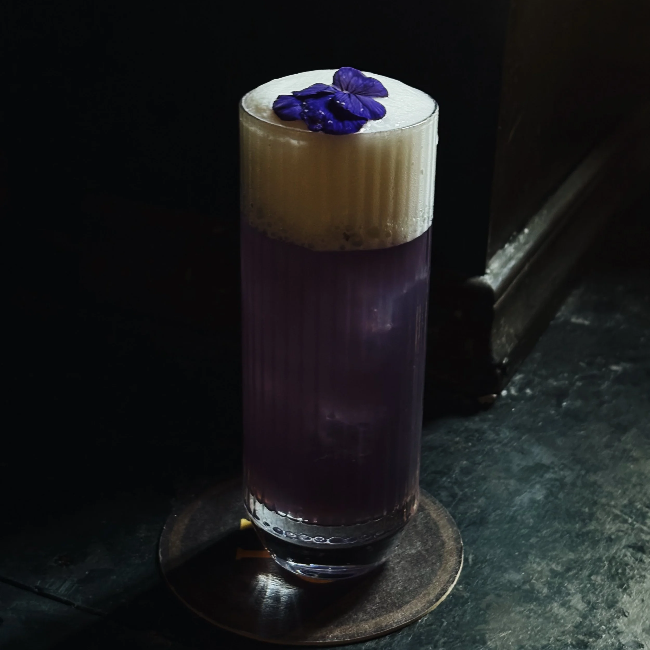 A tall glass filled with a layered beverage topped with purple edible flowers, placed on a dark textured surface.