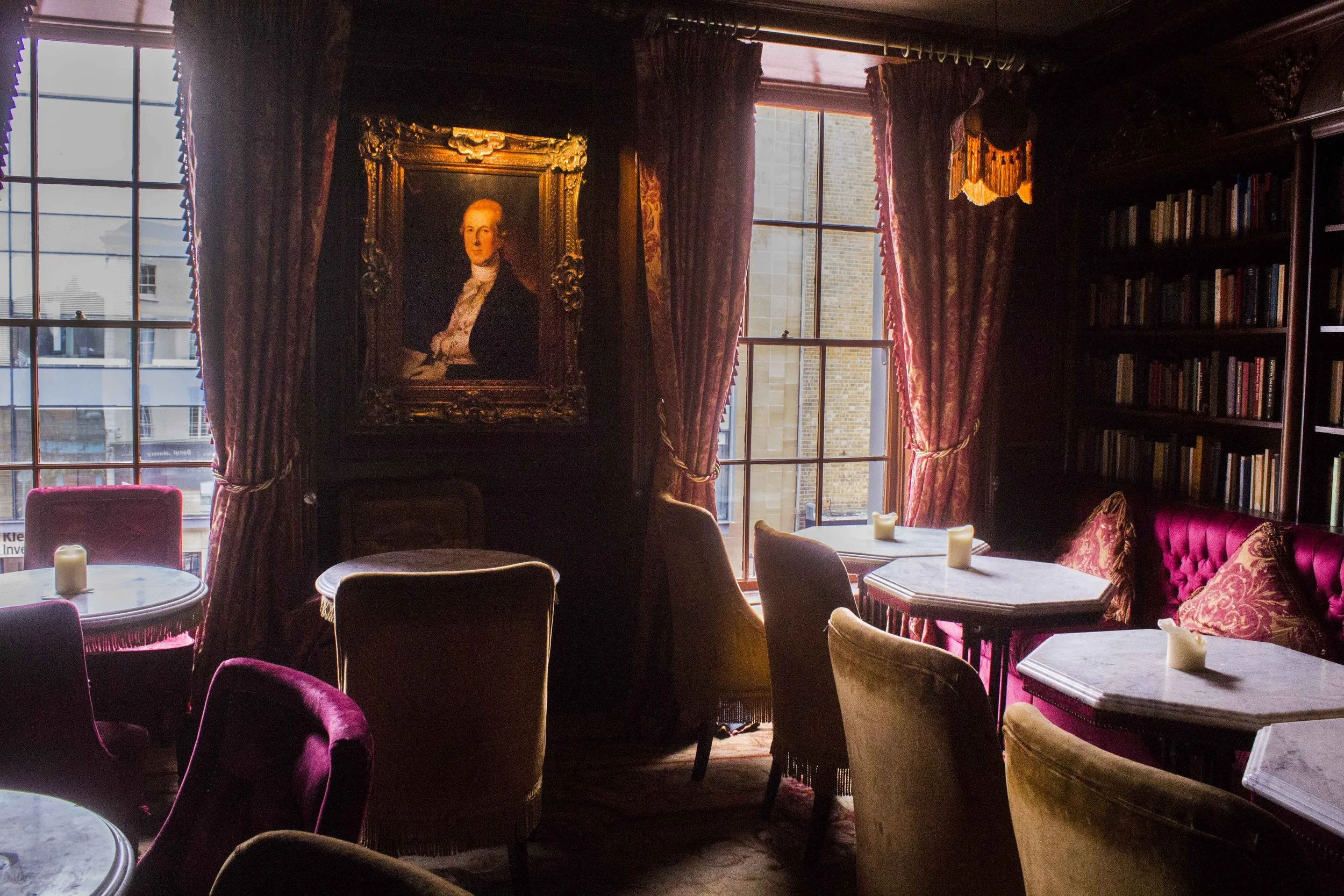 A cozy room with large windows, red velvet chairs, a bookshelf, a framed portrait painting on the wall, and candles on the tables, with natural light coming through the windows.