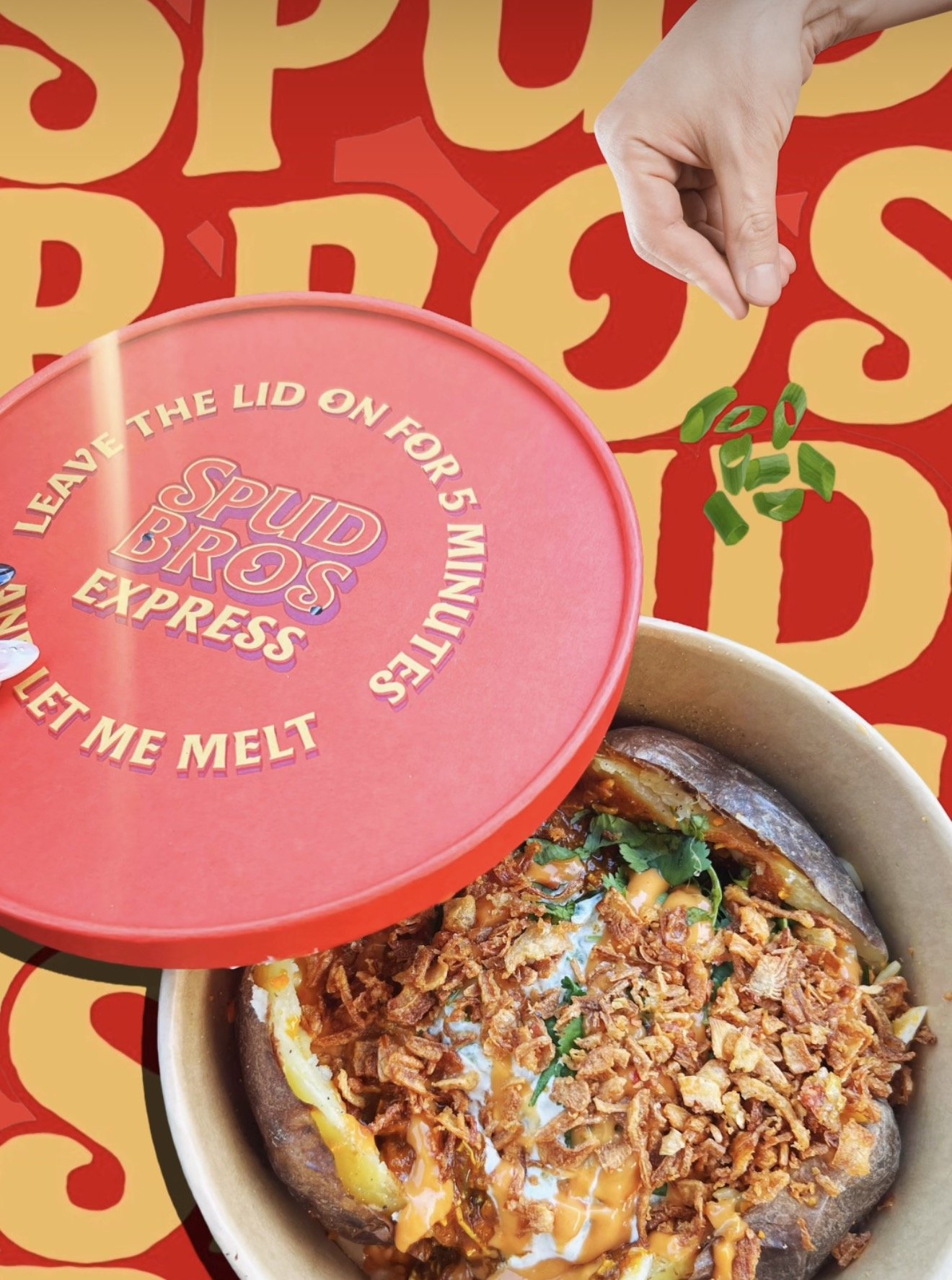 A metal bowl containing a baked potato topped with cheese, sour cream, fried onion bits, and green herbs. There is a red Spud Bros Express lid in the foreground and a colorful background with yellow and red text.