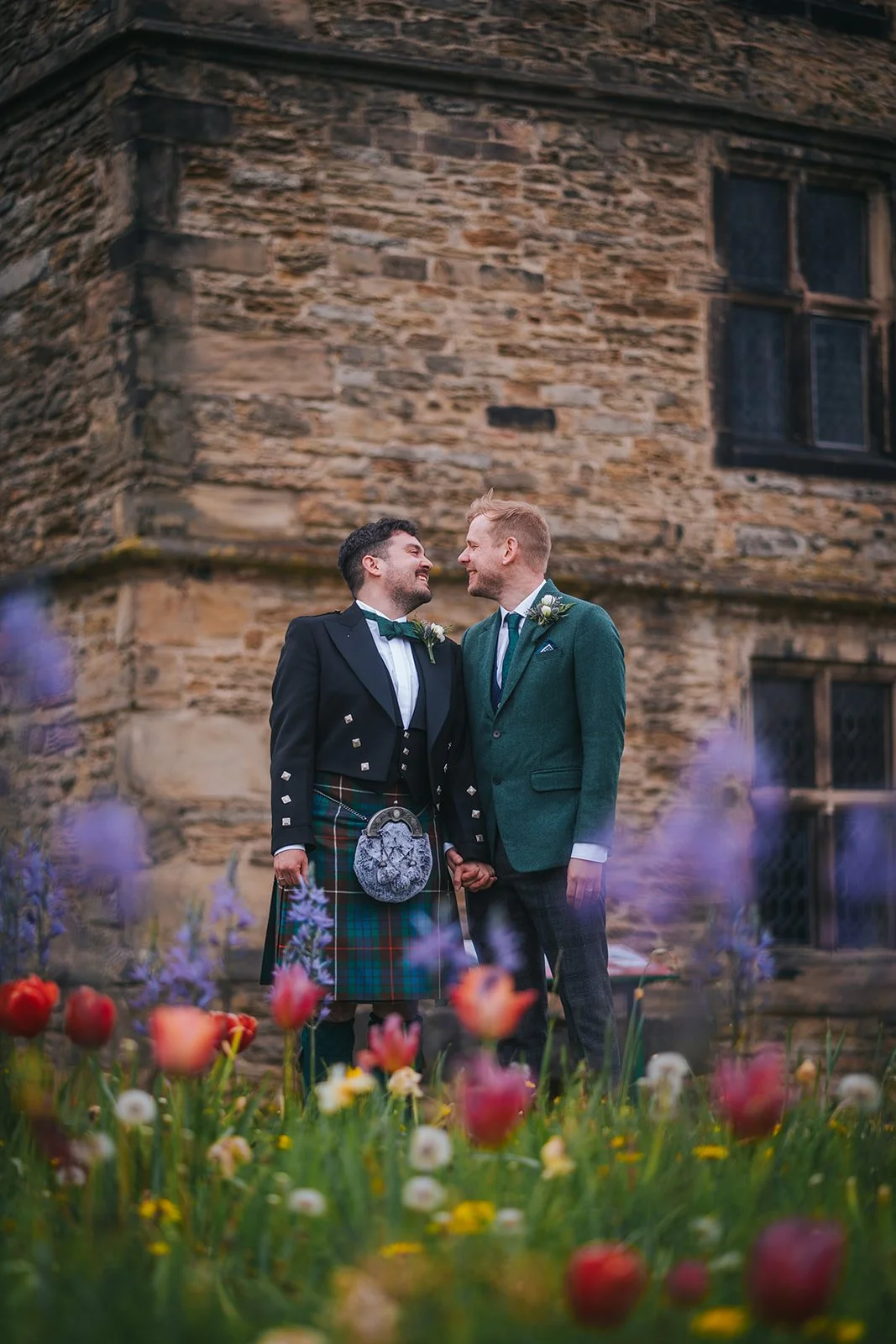 Two grooms, one in a suit with a kilt and the other in a green tweed suit, gaze lovingly at each other in from of a ruin, framed by spring flowers in the foreground